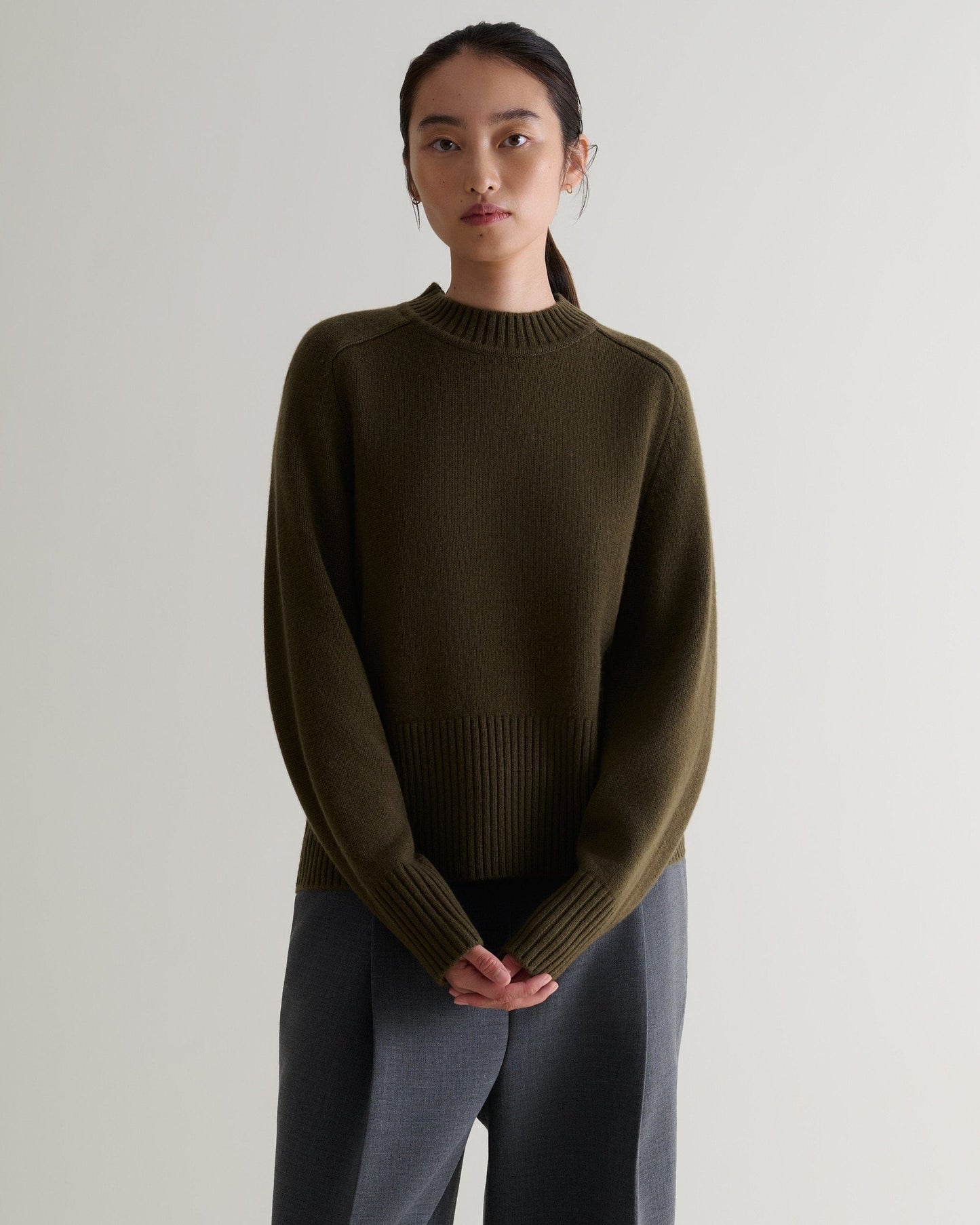 Image 1 of 5 showing woman wearing Rise & Fall's military green cashmere merino saddle sleeve crew neck jumper with grey trousers.