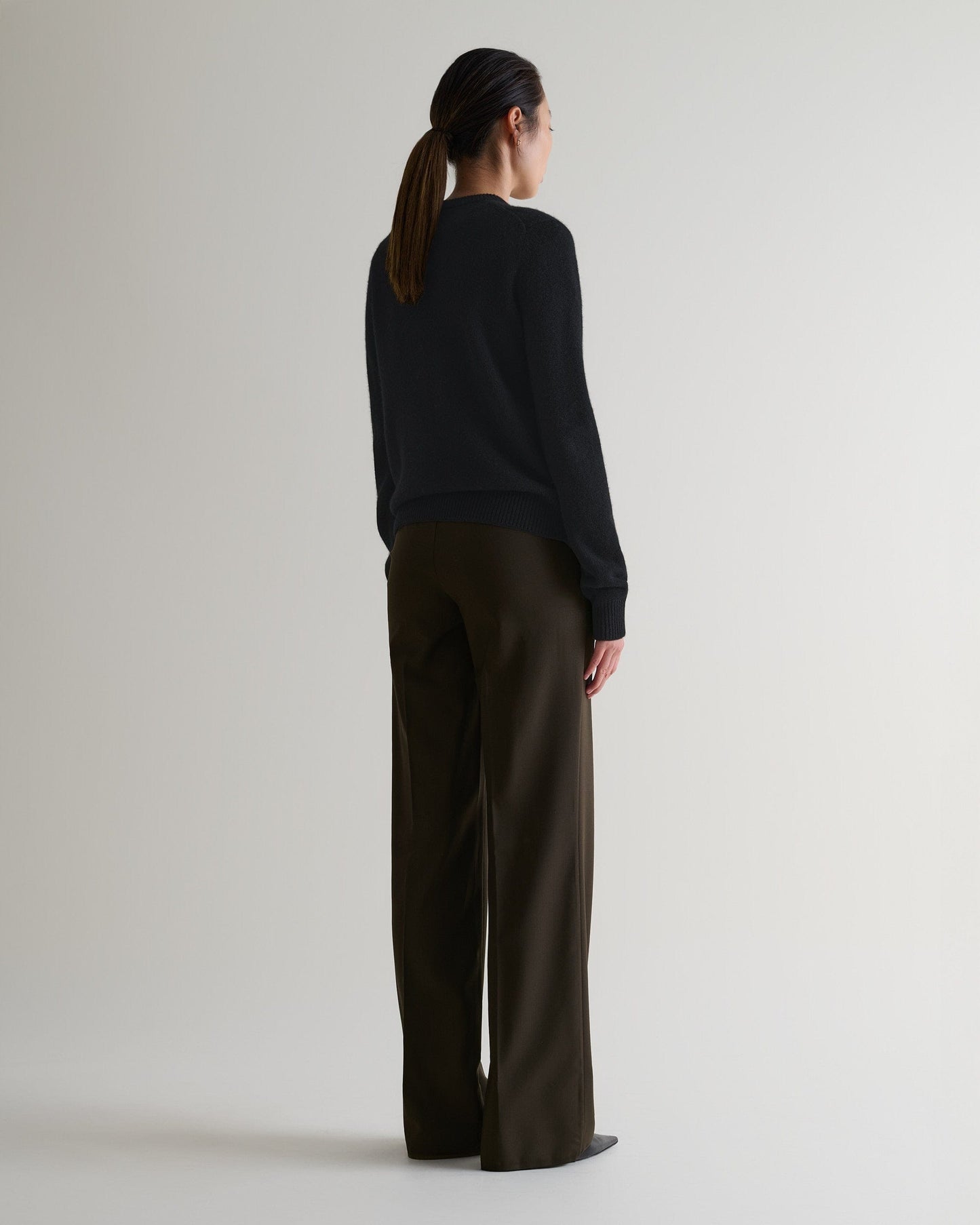 Image 3 of 4 showing the back of a woman wearing Rise & Fall's black mid-weight cashmere crew neck jumper with brown silk trousers.
