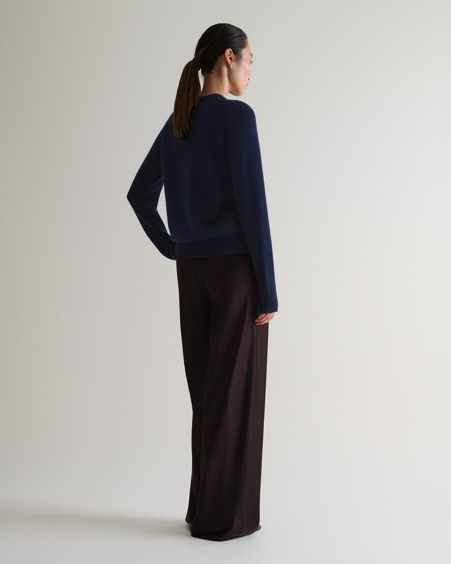 Image 3 of 5 showing the back of a woman wearing Rise & Fall's navy mid weight cashmere crew neck jumper with brown silk trousers.