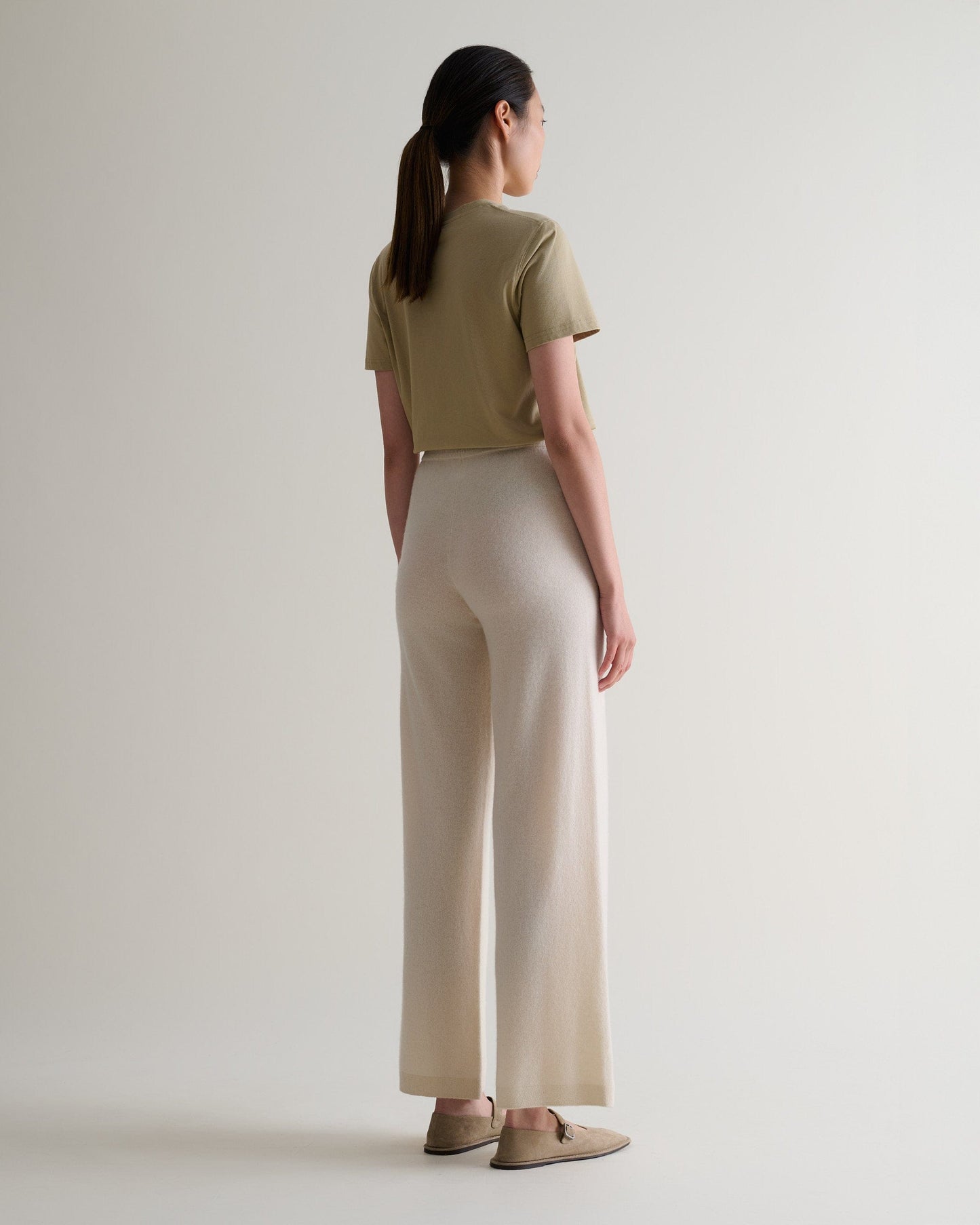 Image 3 of 6 showing back of woman wearing Rise & Fall's cream fine cashmere wide-leg trousers with beige t-shirt and shoes.