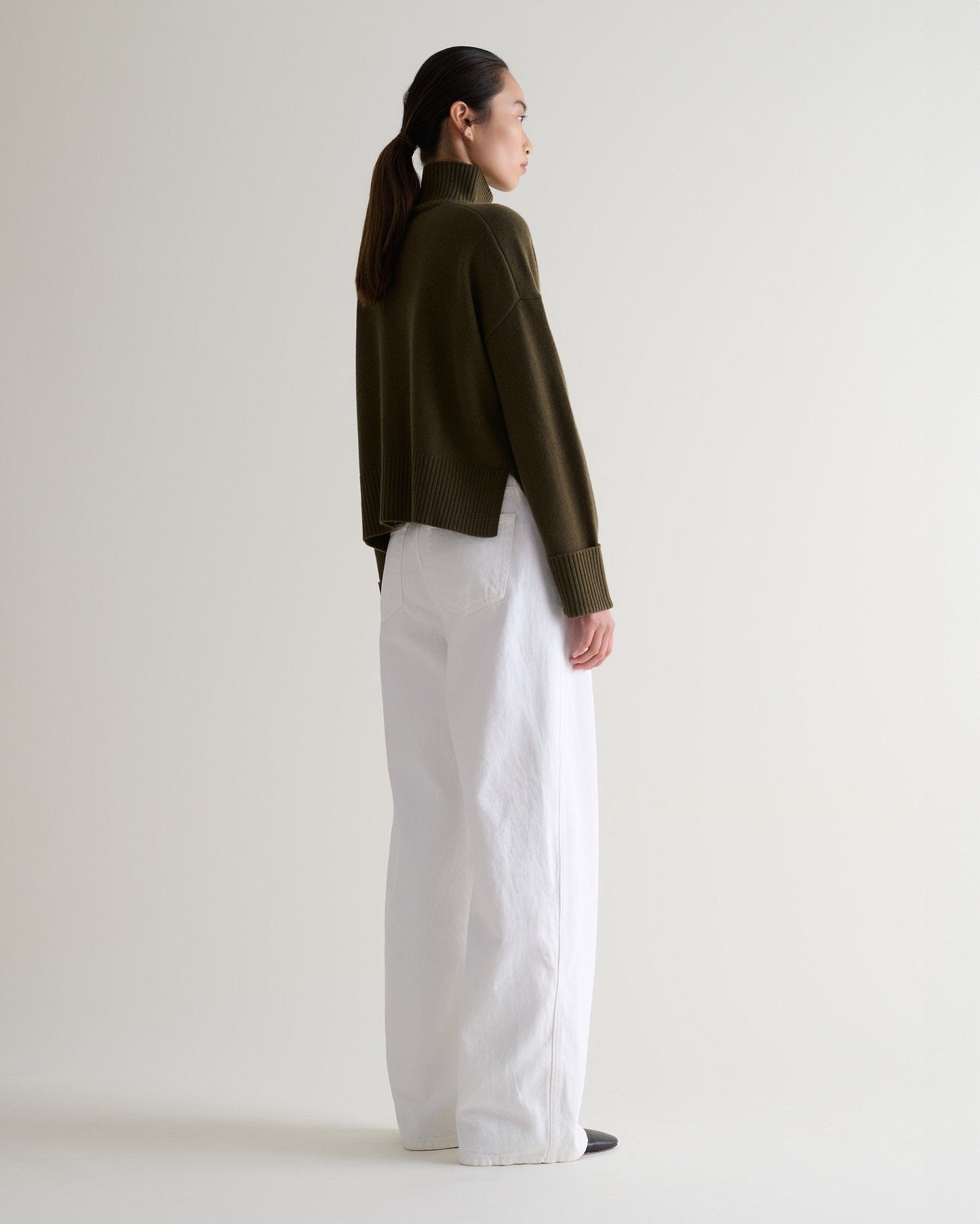 Image 4 of 5 showing back view of a woman wearing Rise & Fall's military green cashmere merino high neck jumper with white jeans.