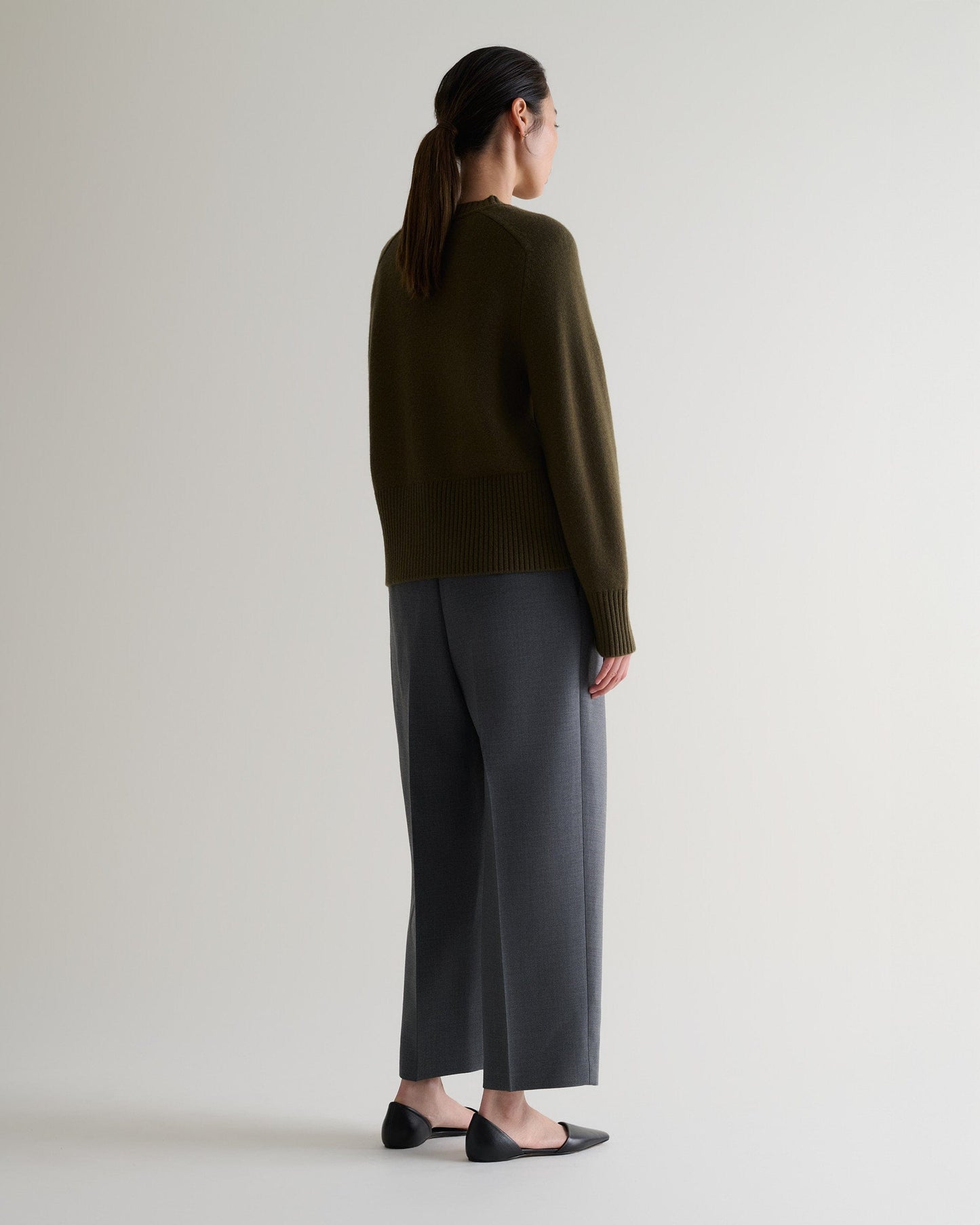Image 4 of 5 showing back view of woman wearing Rise & Fall's military green cashmere merino saddle sleeve crew neck jumper with grey trousers.
