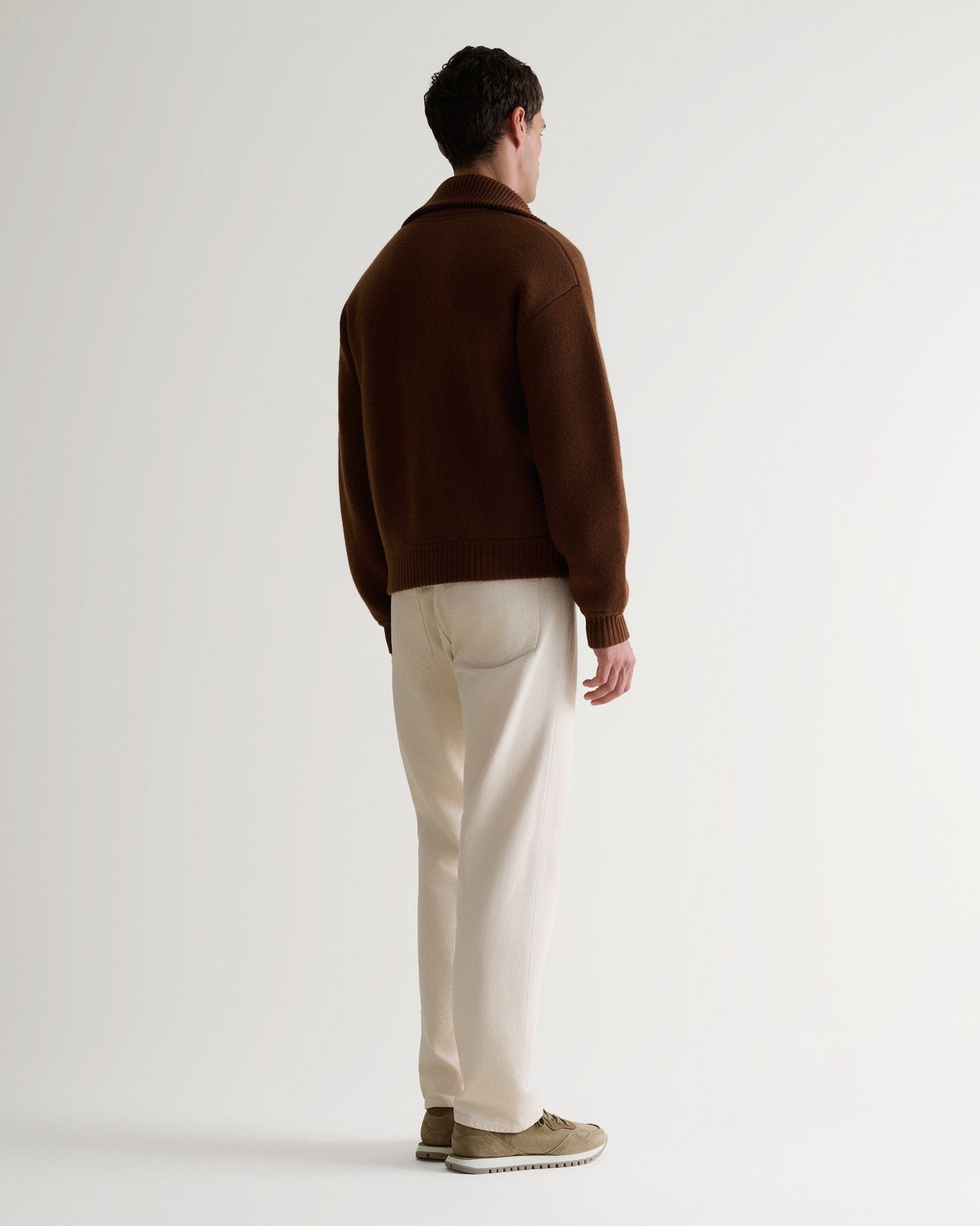 Image 4 of 6 of man wearing Rise & Fall's burl brown cashmere merino stand neck ribbed jumper with cream jeans and beige suede trainers. Full length back view.