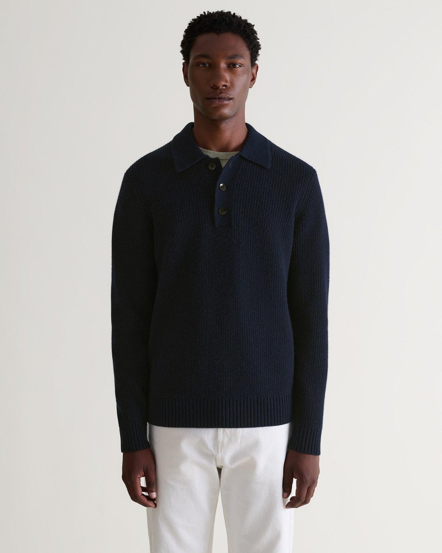 Image 4 of 6 showing man wearing Rise & Fall's navy cashmere merino fisherman ribbed polo sweater. Worn with green T-shirt and white jeans.