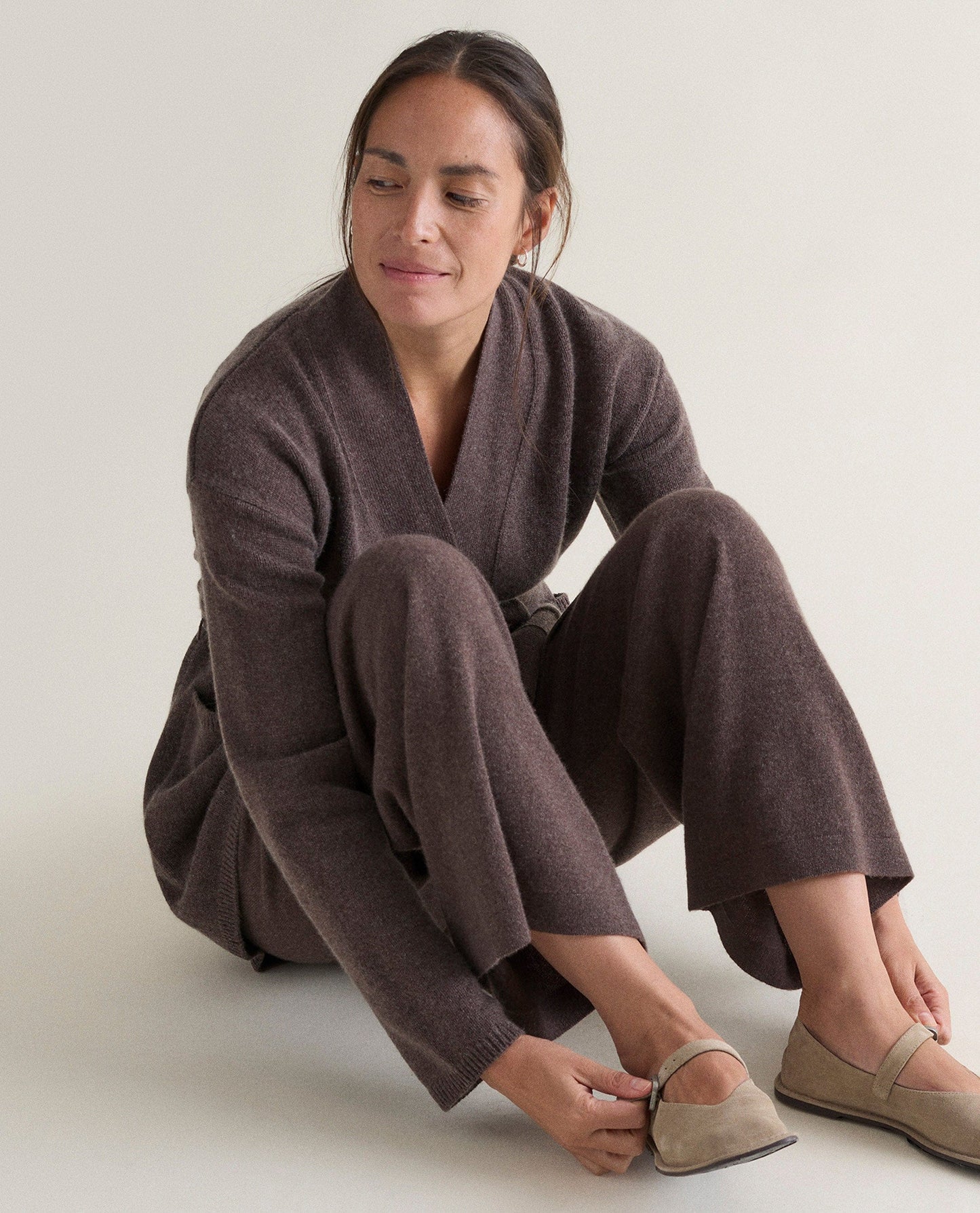 Image 4 of 6 showing woman sat on floor wearing Rise & Fall's brown cashmere merino belted cardigan with matching cashmere trousers and light suede shoes.
