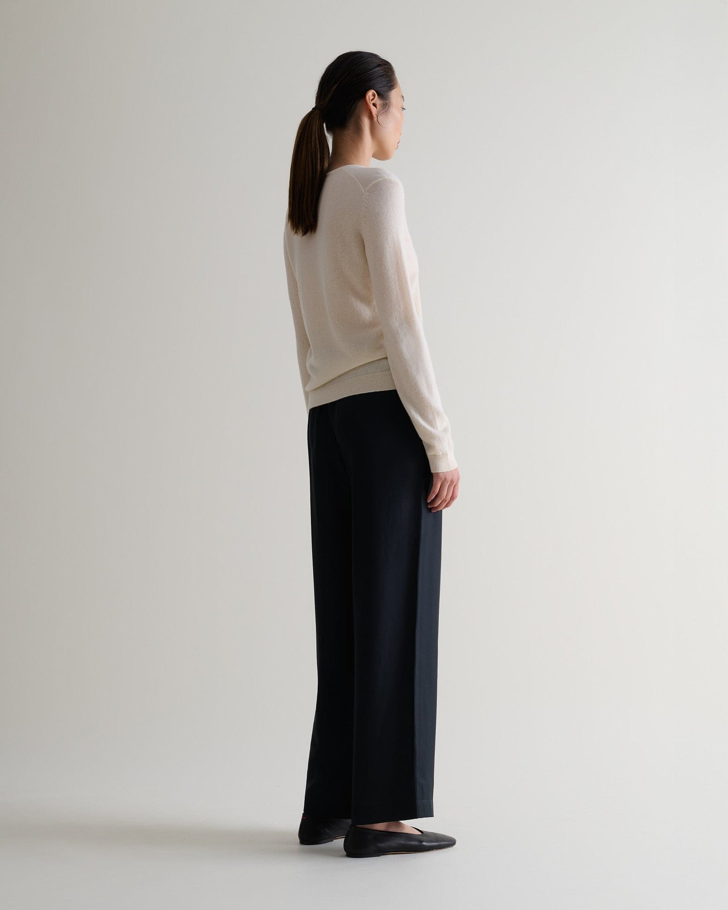 Image 5 of 5 showing side view of a woman wearing Rise & Fall's buttermilk cashmere merino henley jumper with black silk trousers.
