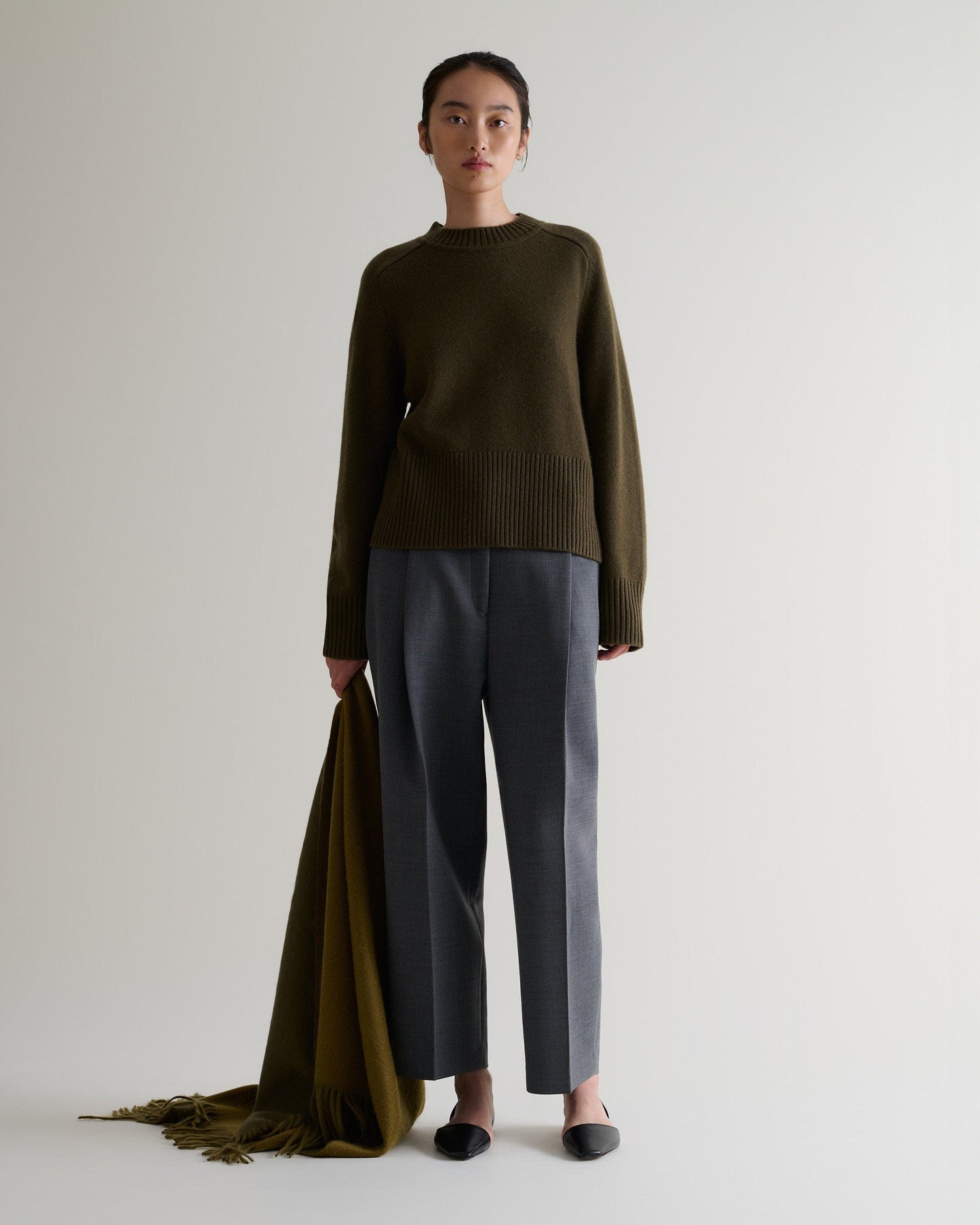 Image 5 of 5 showing woman wearing Rise & Fall's military green cashmere merino saddle sleeve crew neck jumper with grey trousers, holding olive cashmere wrap.