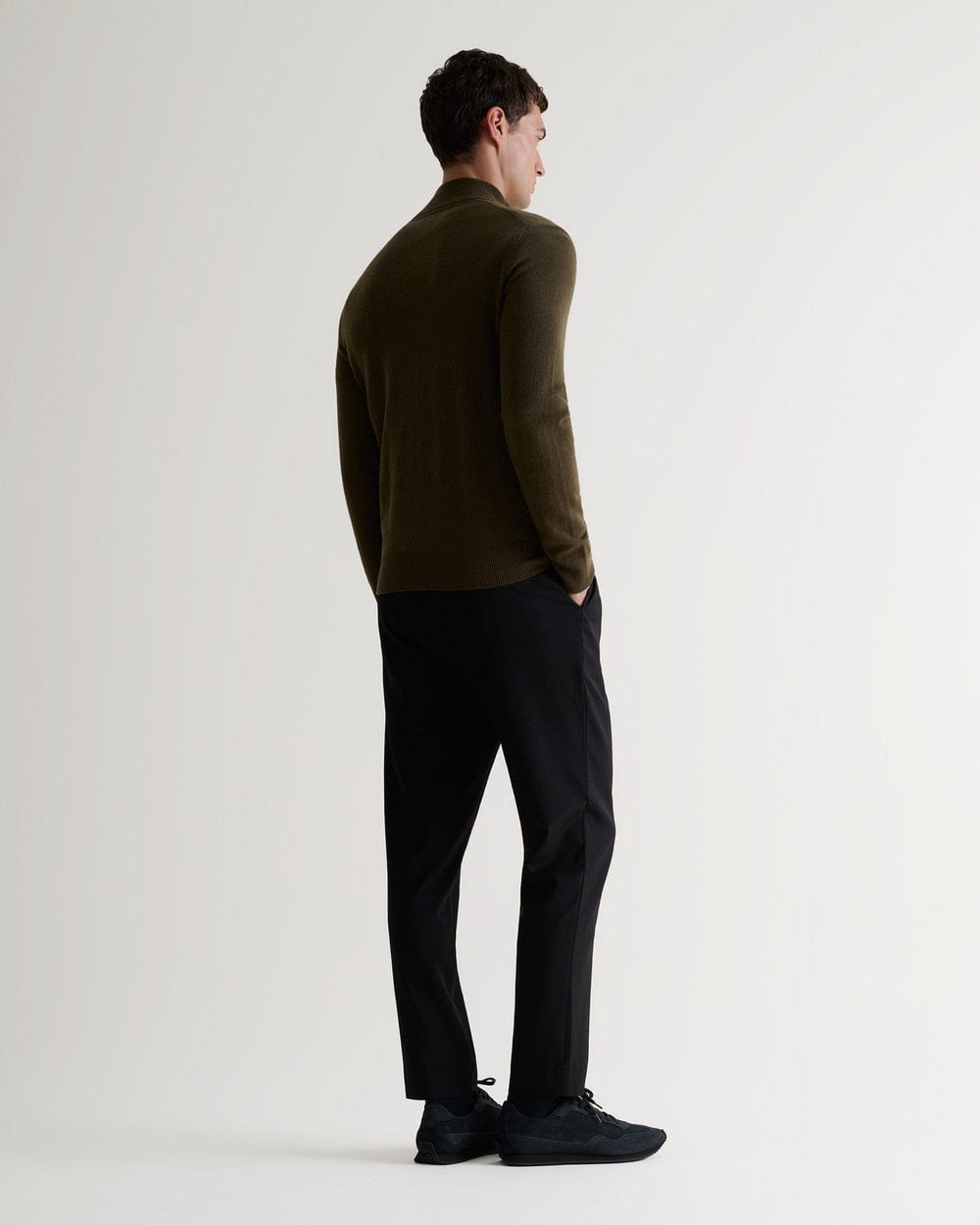 Full length back view of man wearing Rise & Fall's military green finest cashmere 1/4 zip jumper. Worn with black trousers.