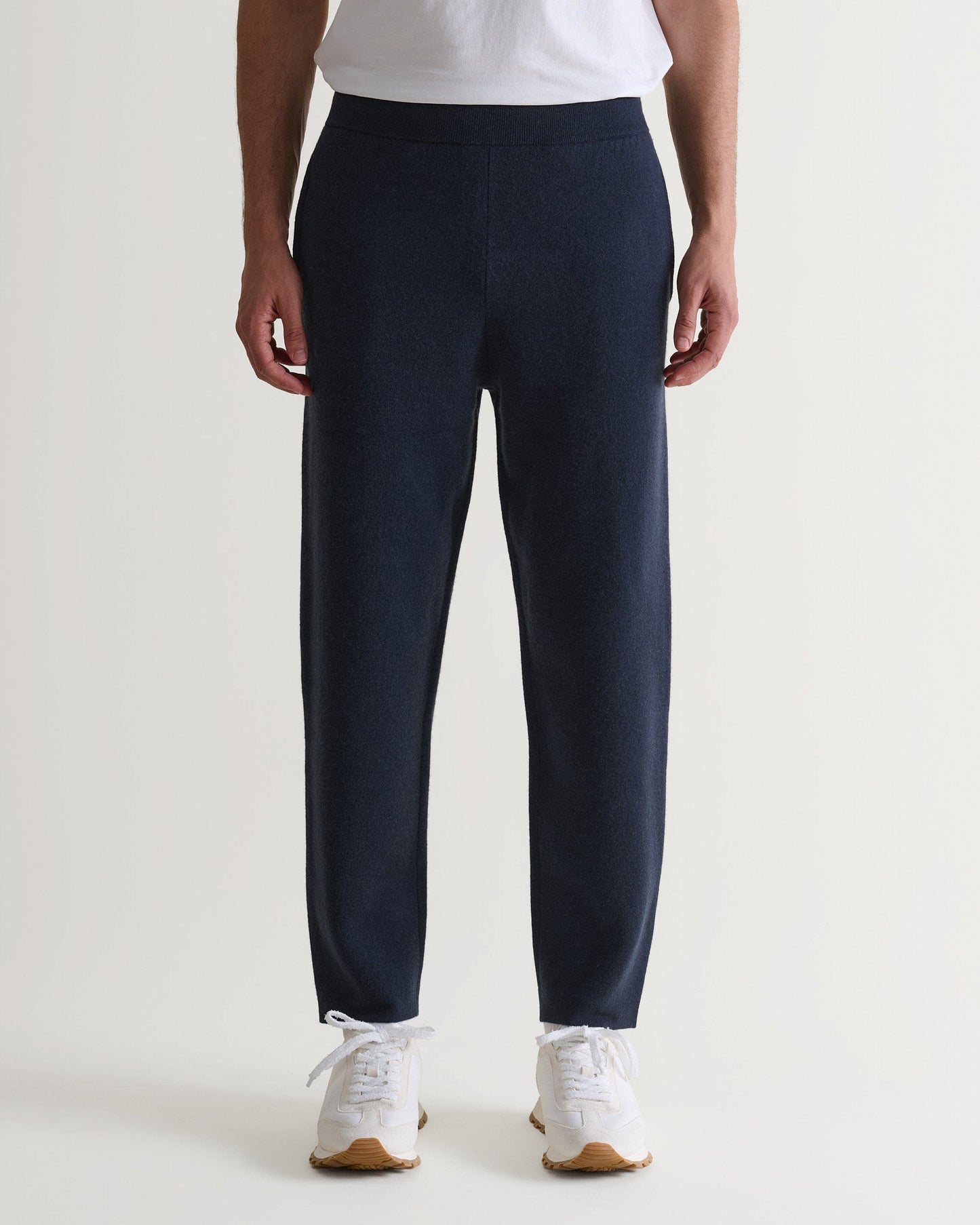 Bottom half of man wearing Rise & Fall's navy cashmere merino double-faced joggers with white T-shirt and trainers.