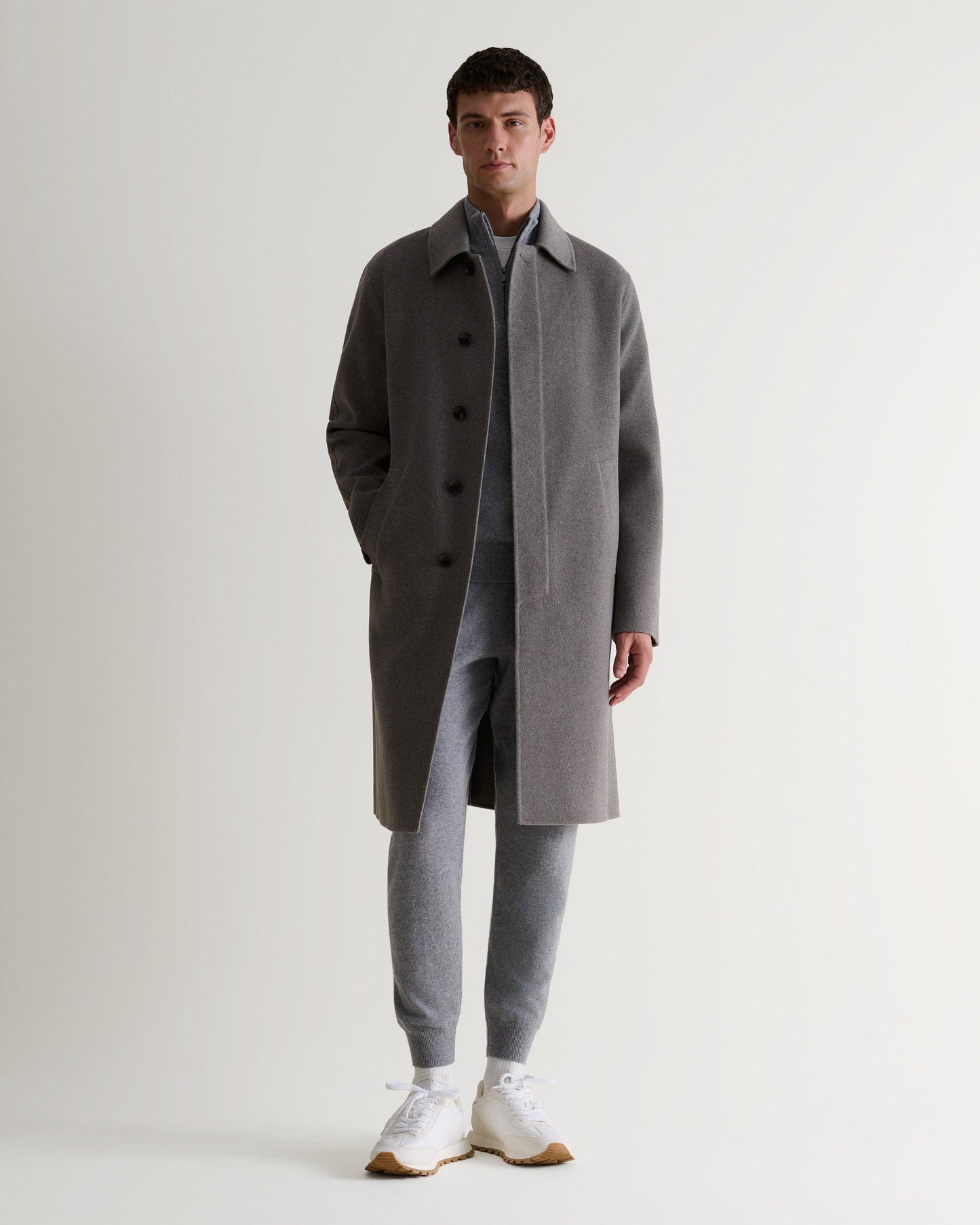 Man wears fawn cashmere merino double-faced wool coat. Matched with grey cashmere jumper and joggers. Full length view.