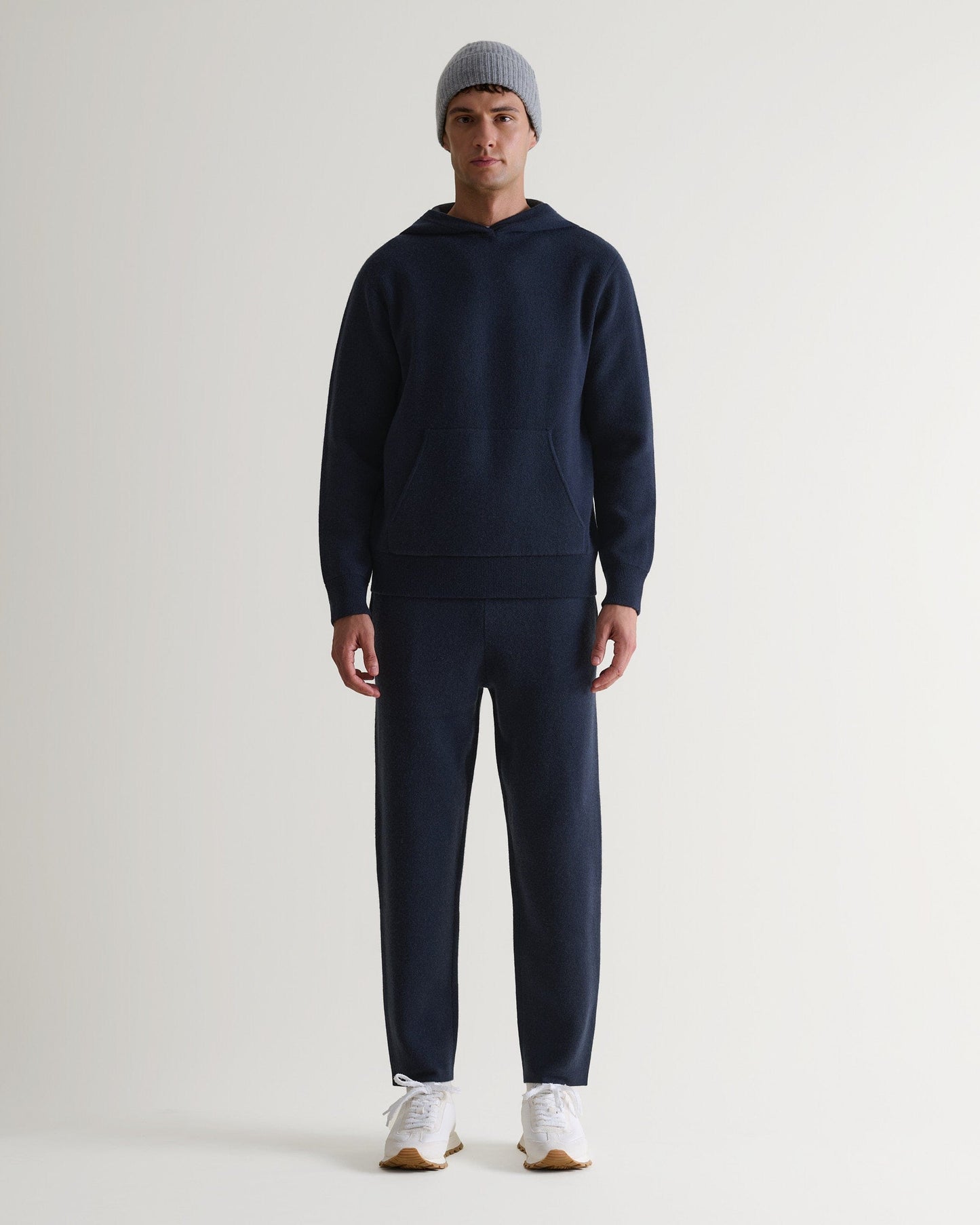 Man wears navy cashmere merino double-faced knitted hoodie with matching trousers, white trainers and light grey beanie.