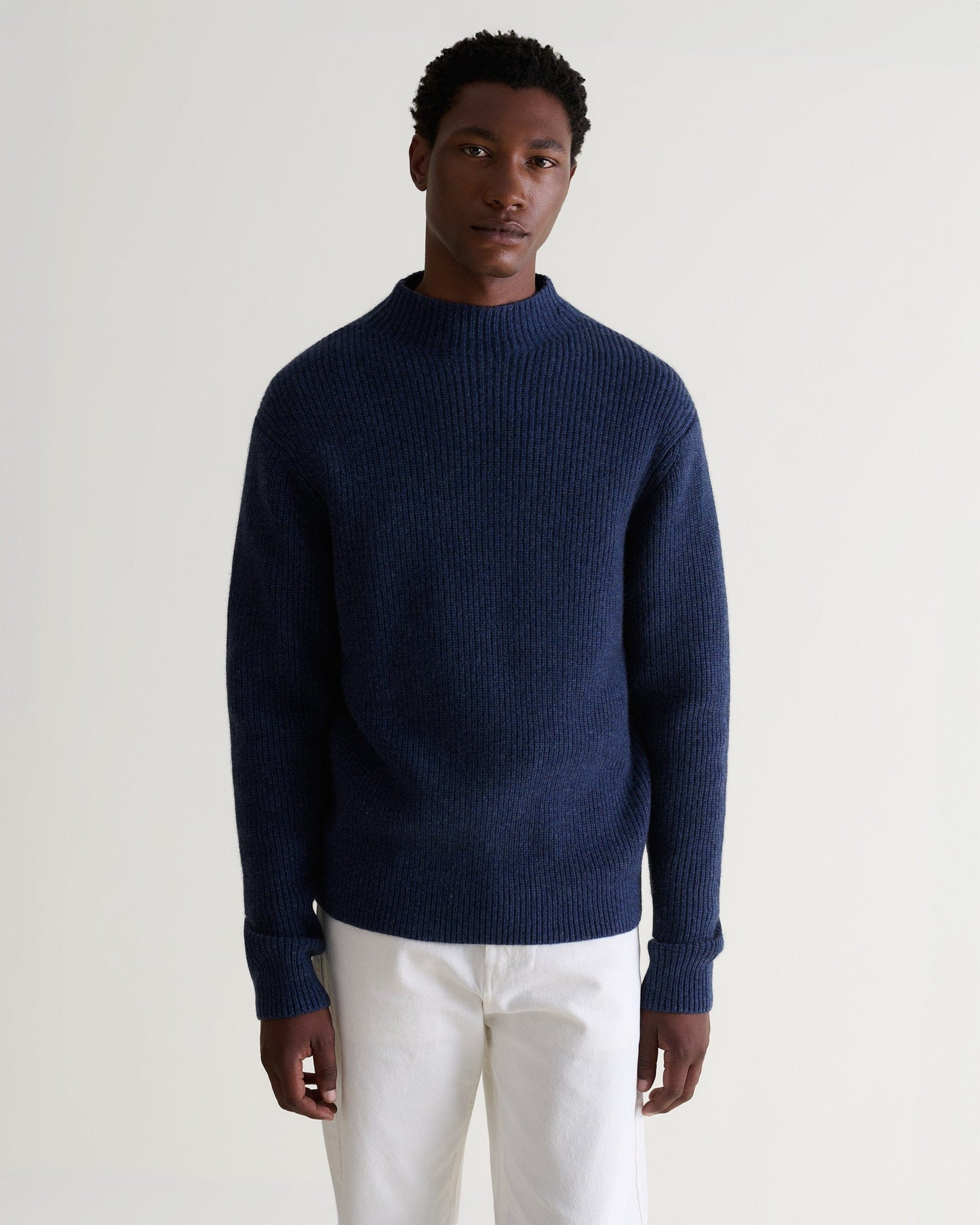 Front view of man wearing Rise & Fall's indigo blue cashmere merino stand neck jumper with white trousers.