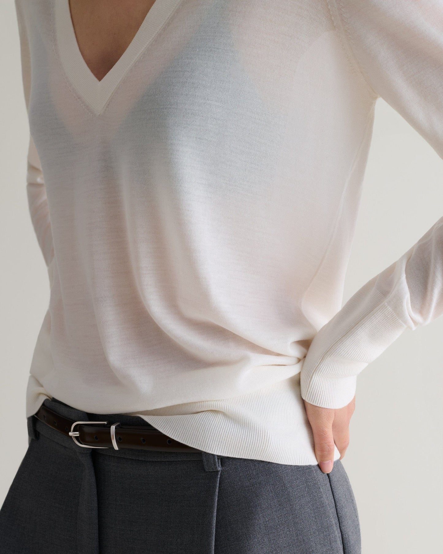 Close up of woman wearing Rise & Fall's cream superfine merino wool v-neck jumper. Worn with grey trousers and a black belt.