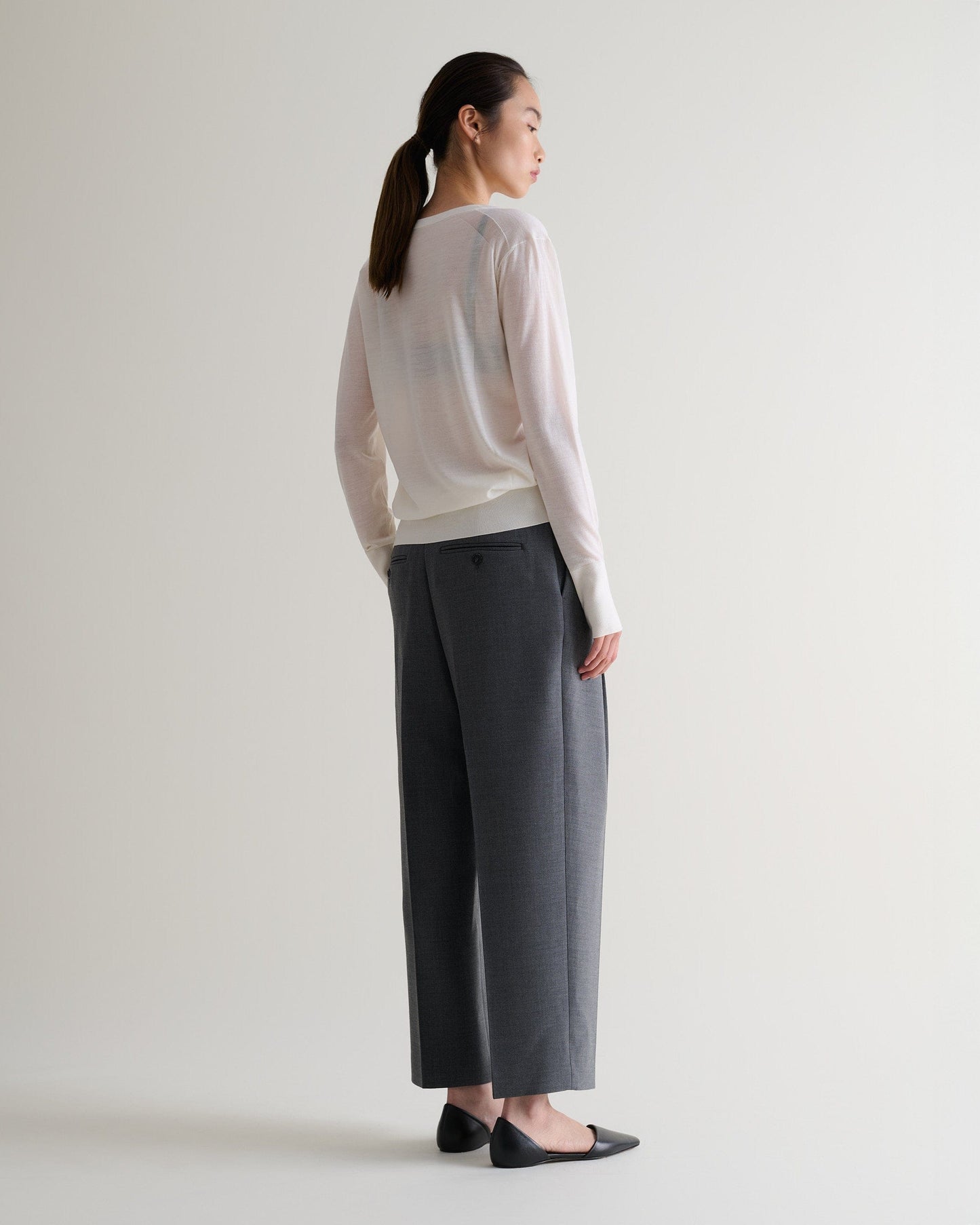 Full length back view of woman wearing Rise & Fall's cream superfine merino wool v-neck jumper. Worn with grey trousers.