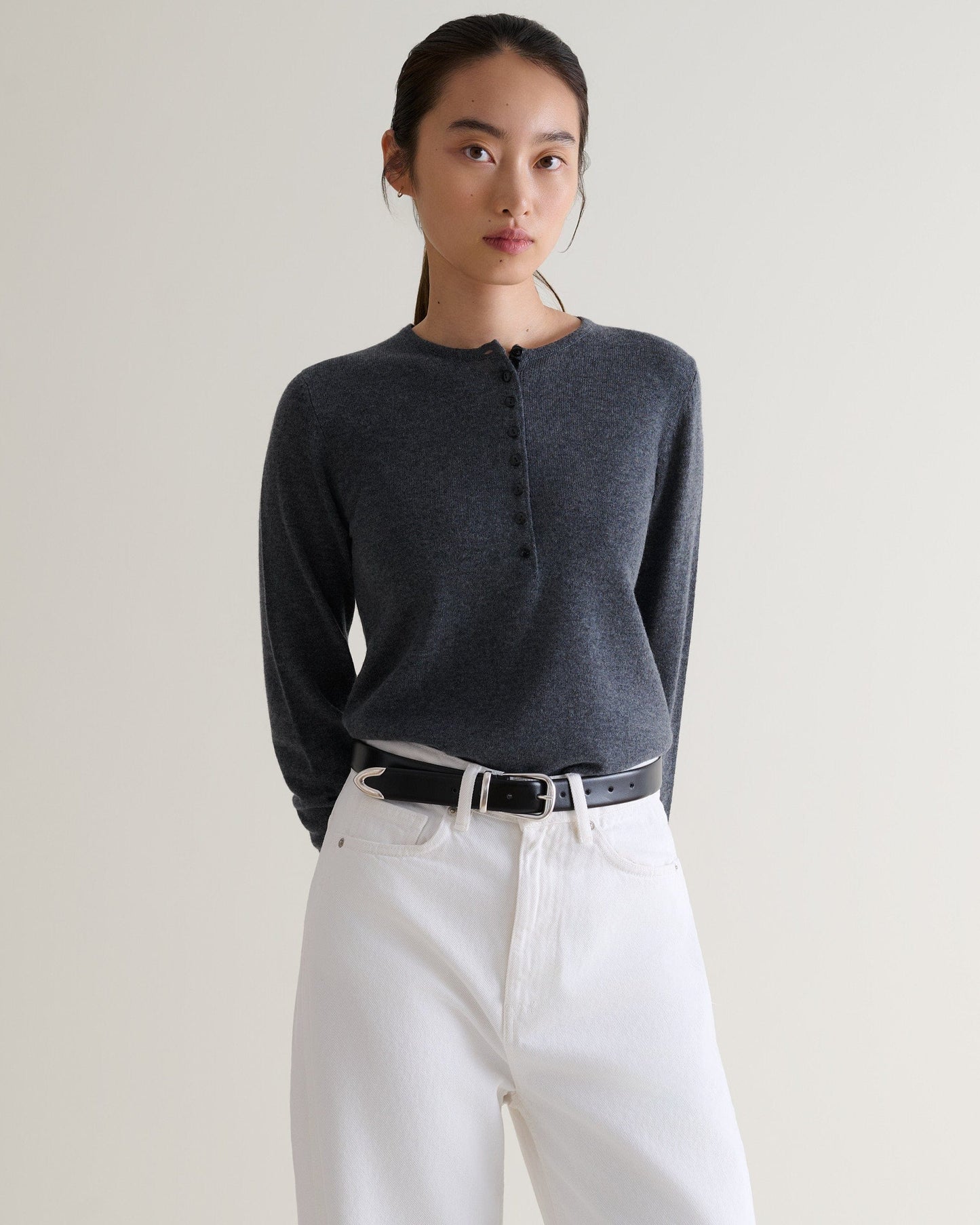 Front view of woman wearing Rise & Fall's graphite cashmere merino henley jumper. Worn with white jeans and black belt.