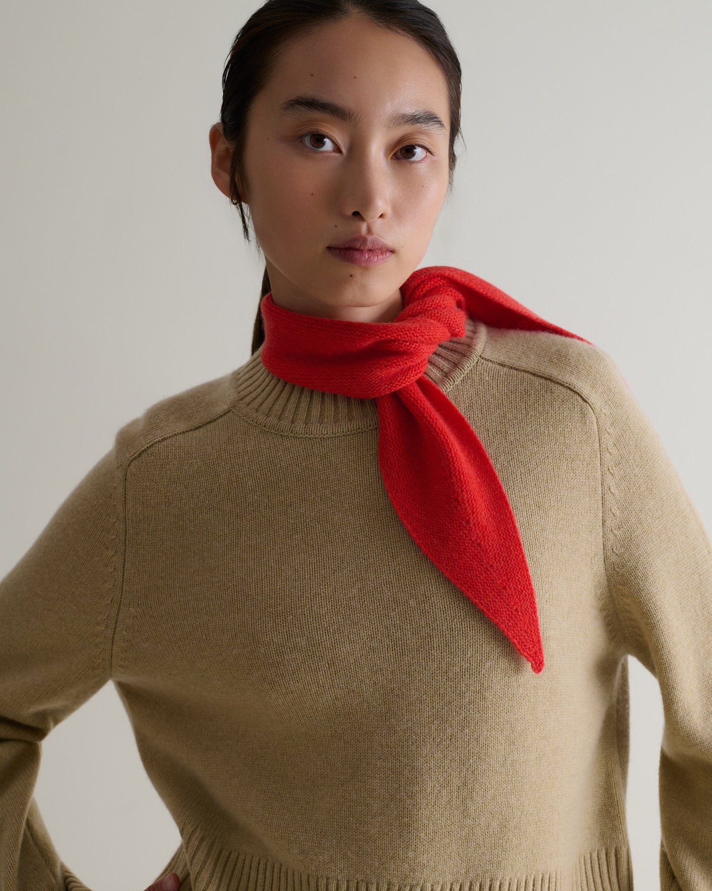 Woman wears Rise & Fall's poppy red finest cashmere ribbed micro bandana. Worn to the side. Paired with beige jumper.
