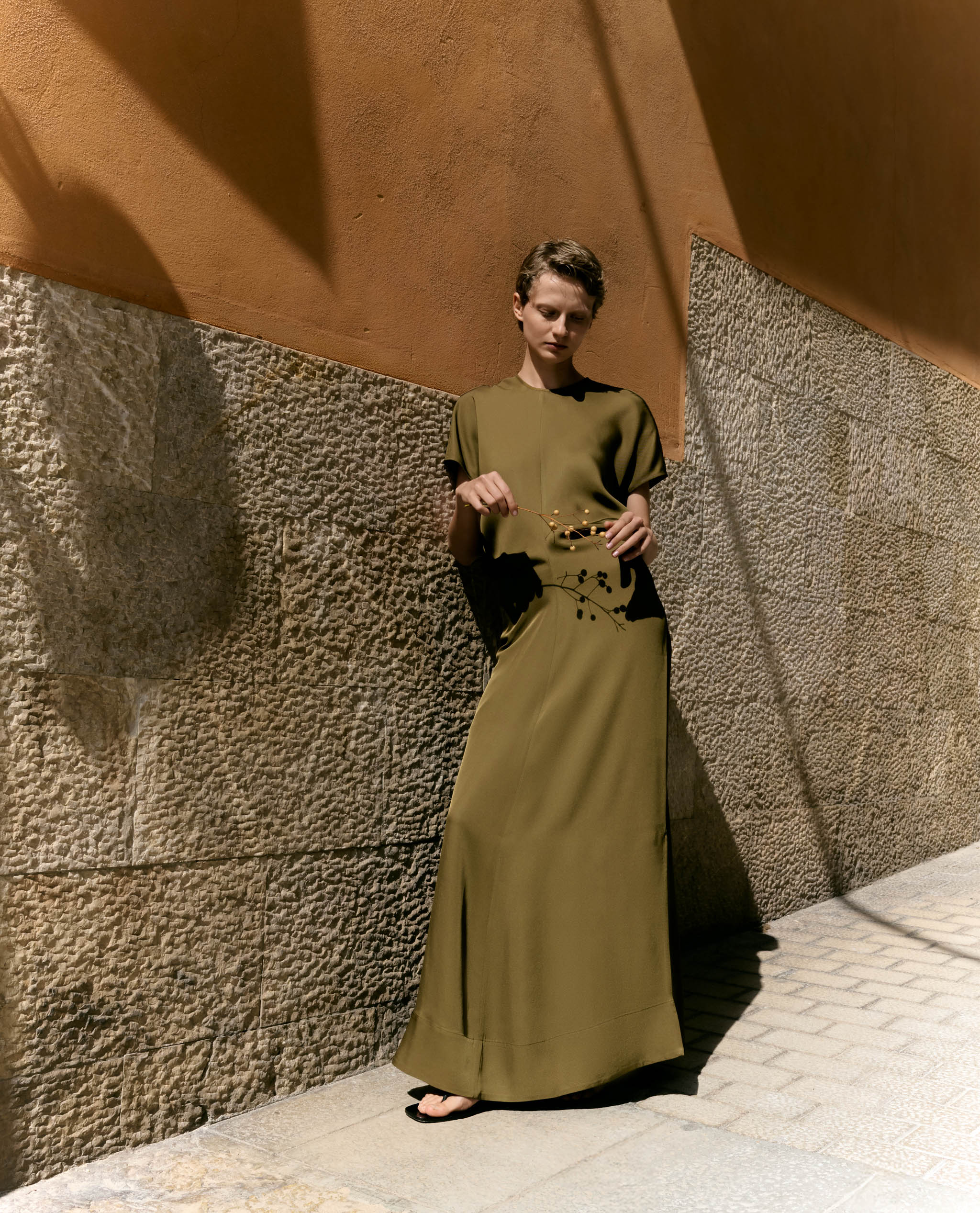 Woman leans against a stone wall while she models an olive Naia maxi dress.