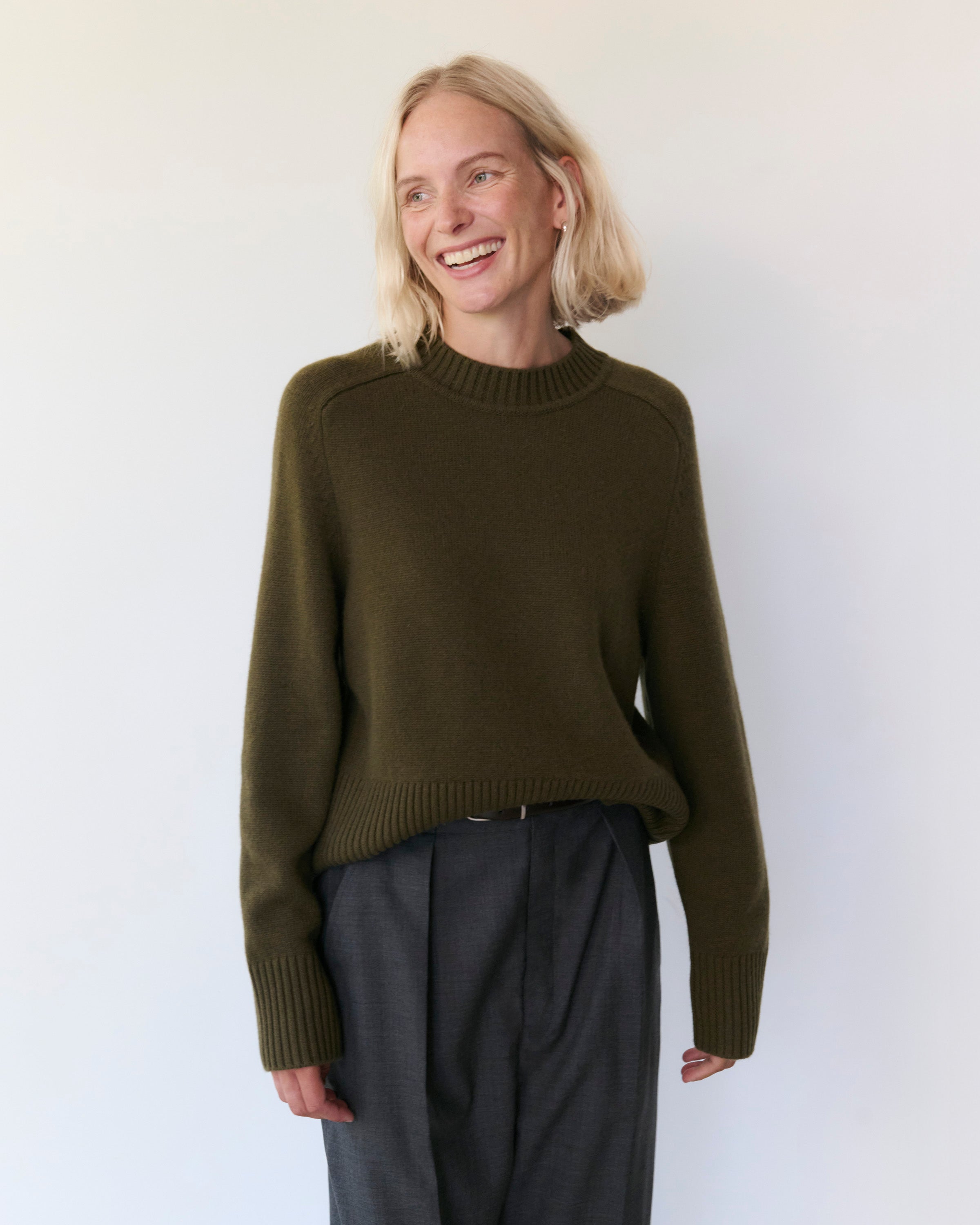 Woman wears Rise & Fall's khaki cashmere crew neck jumper with grey trousers.