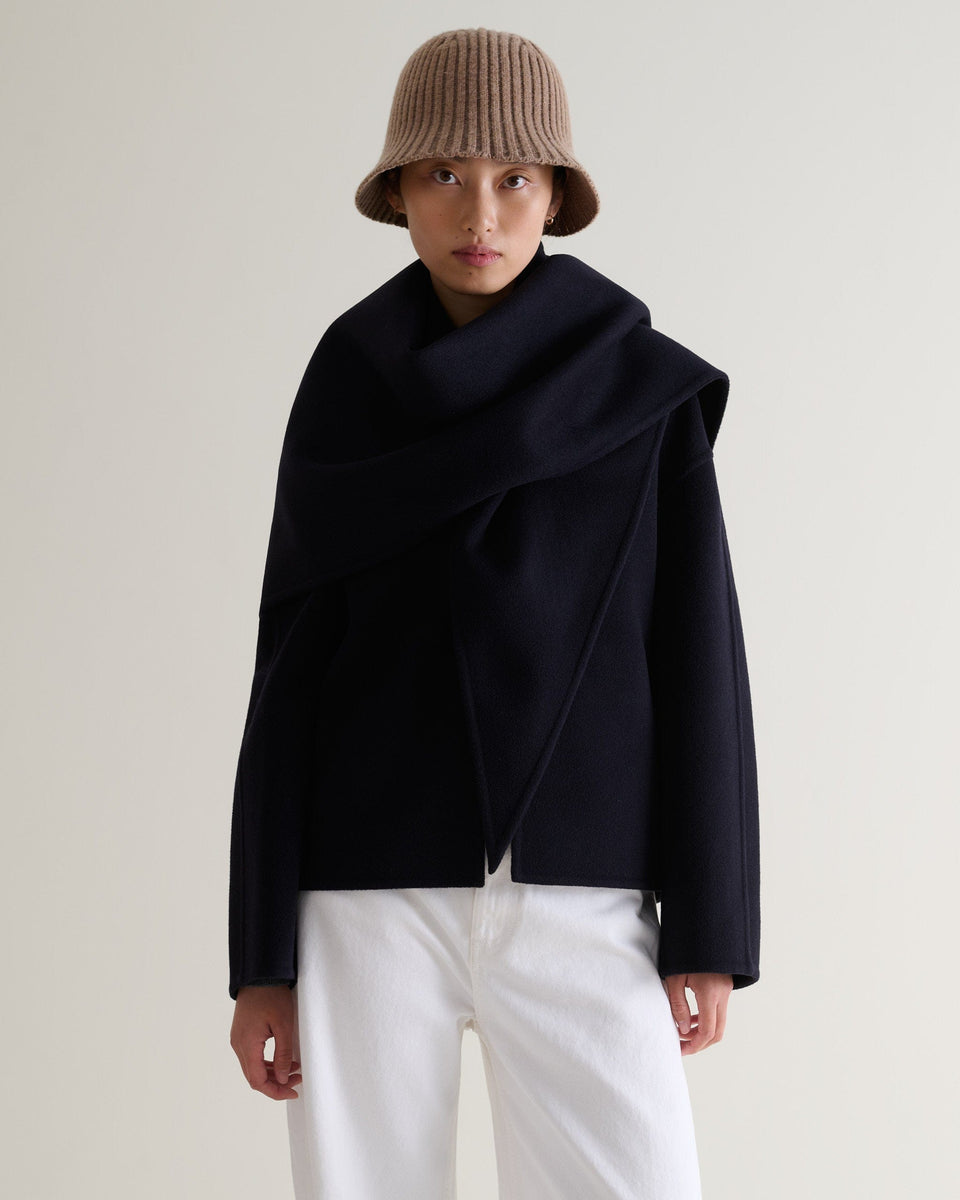 Front view of woman wearing beige merino wool ribbed bucket hat with white trousers and navy cashmere wool scarf jacket.