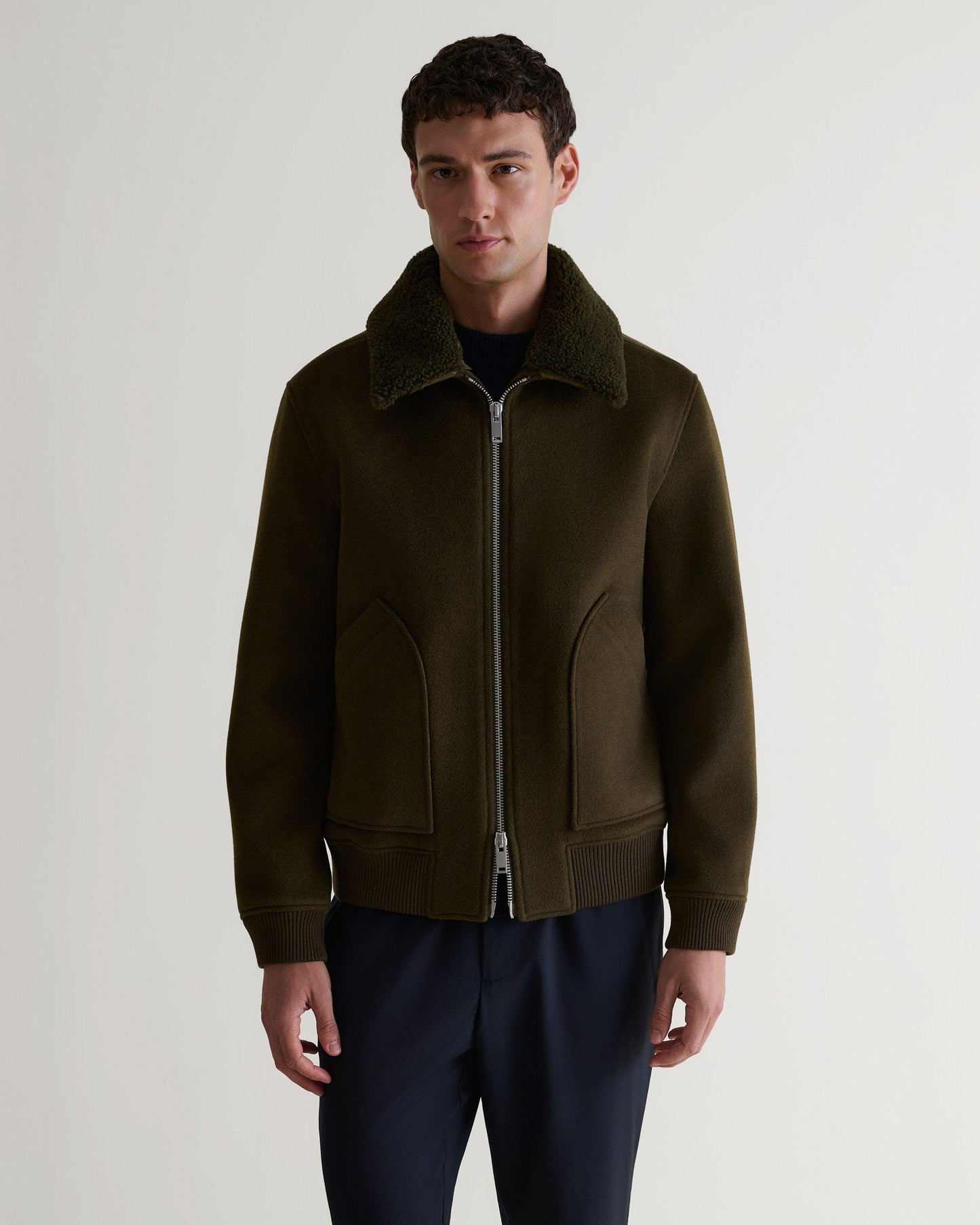 Man wears Rise & Fall's olive wool bomber jacket with shearling collar. Closed zip.