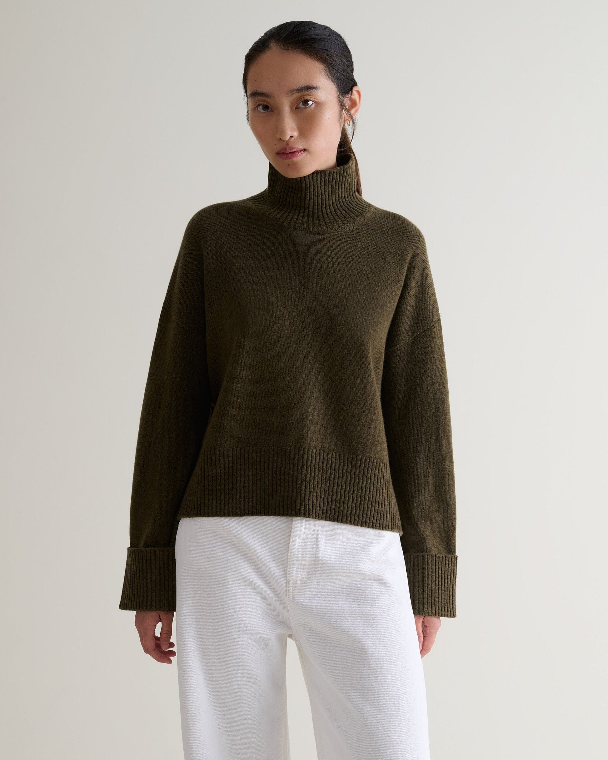 Image 1 of 5 showing woman wearing Rise & Fall's military green cashmere merino high neck jumper with white jeans. 