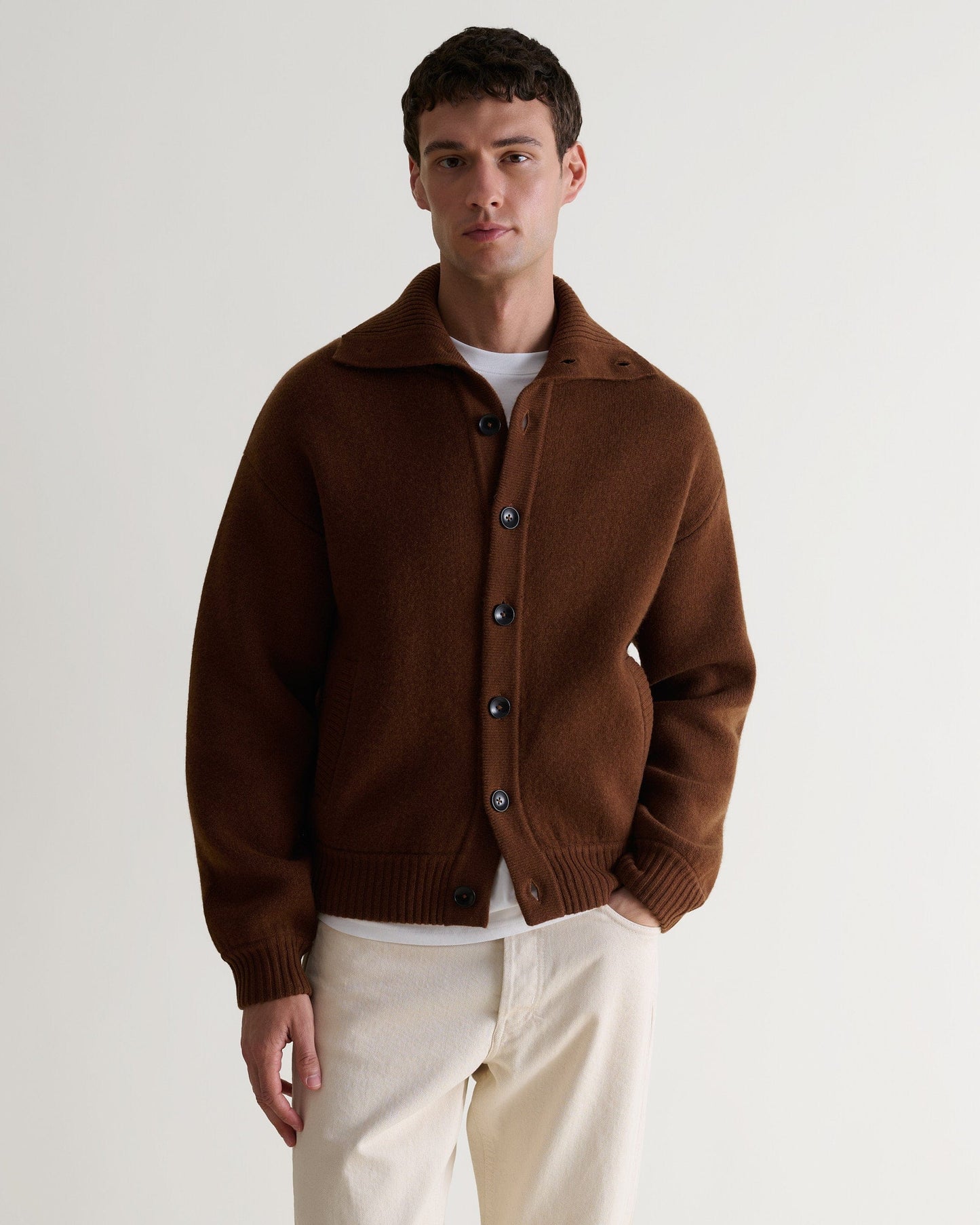 Image 1 of 6 showing man wearing Rise & Fall's burl brown cashmere merino buttoned knitted bomber. Worn with white T-shirt and cream trousers.