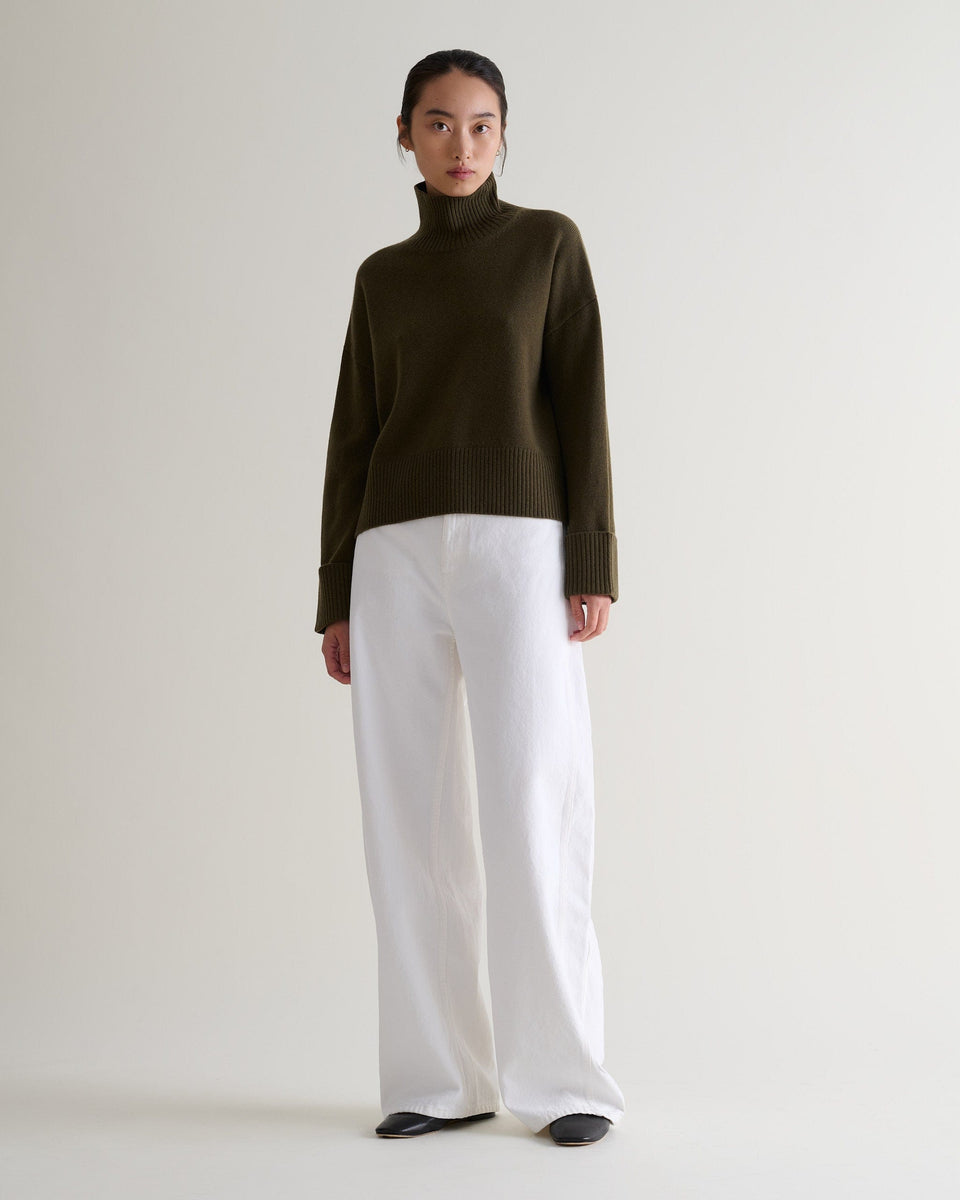 Image 3 of 5 showing woman wearing Rise & Fall's military green cashmere merino high neck jumper with white jeans. 