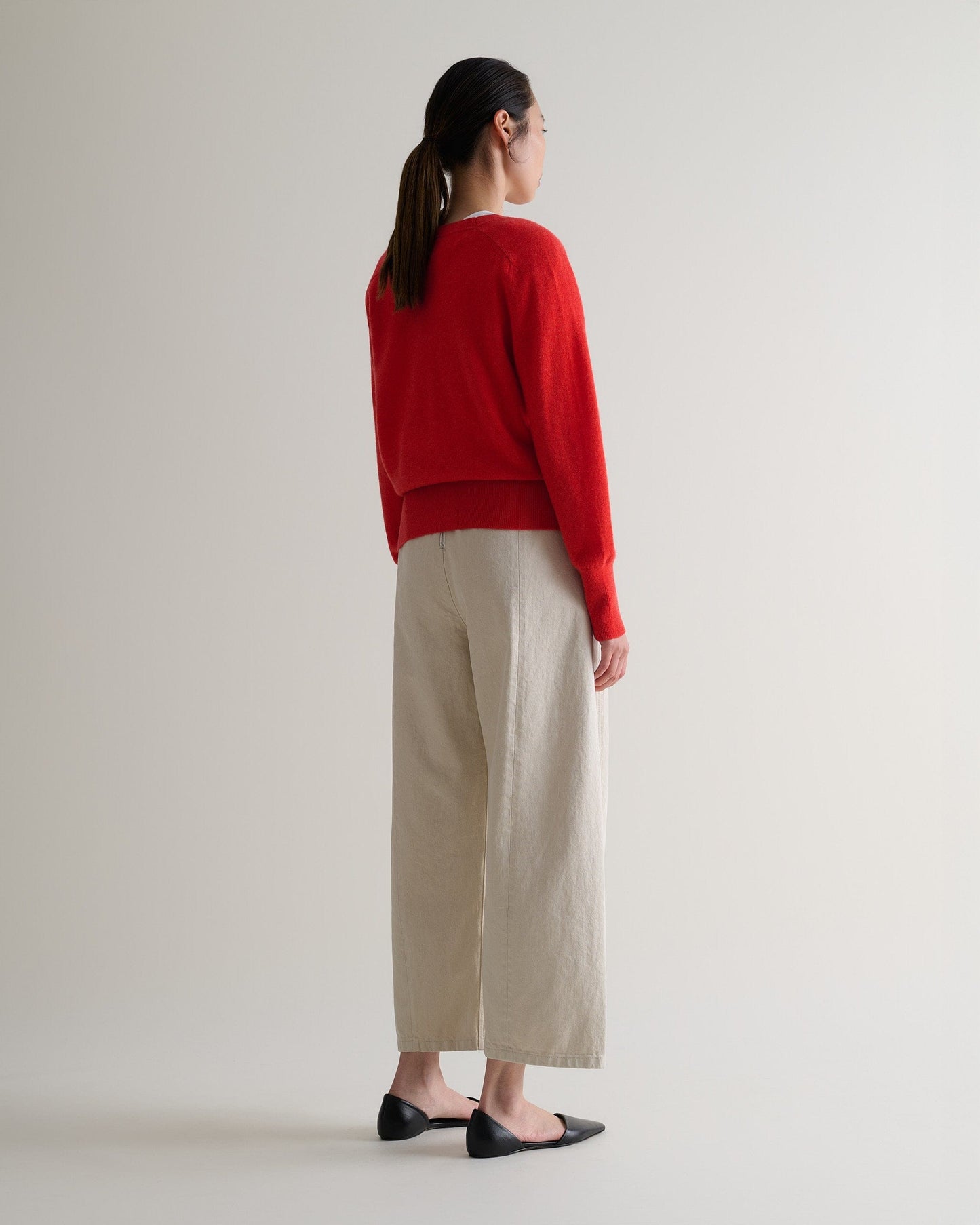 Image 3 of 5 showing back of woman wearing Rise & Fall's poppy red fine cashmere blouson cardigan with cream trousers.