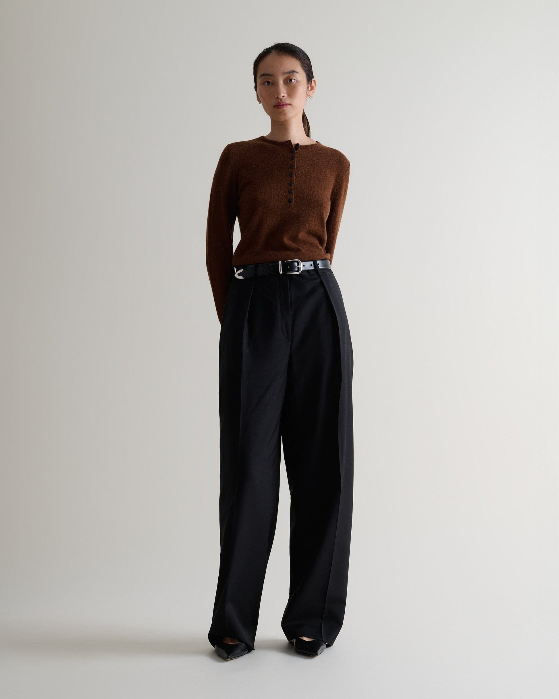Image 3 of 5 showing a woman wearing Rise & Fall's brown cashmere merino henley jumper with black trousers and a black belt. 