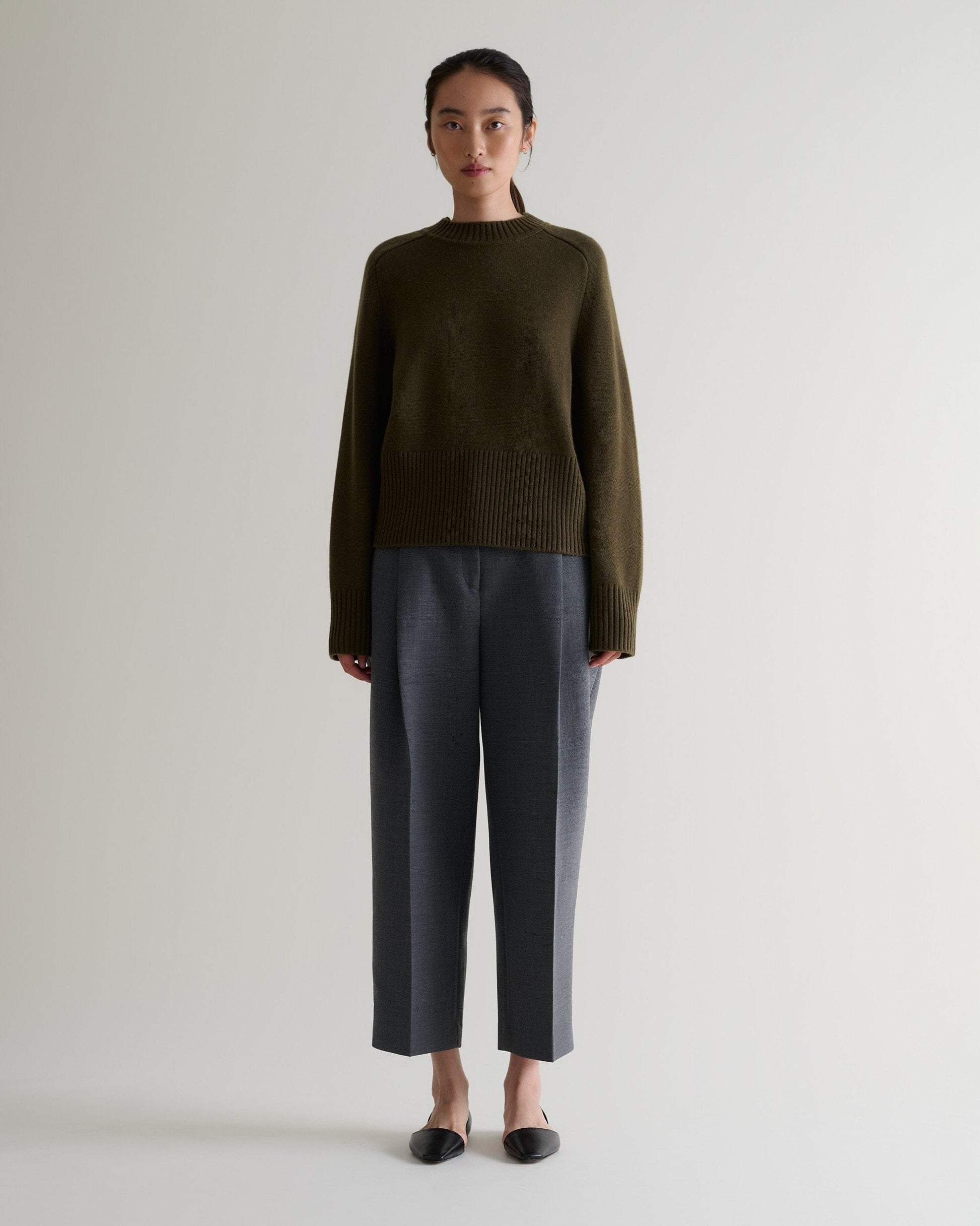 Image 3 of 5 showing woman wearing Rise & Fall's military green cashmere merino saddle sleeve crew neck jumper with grey trousers. 