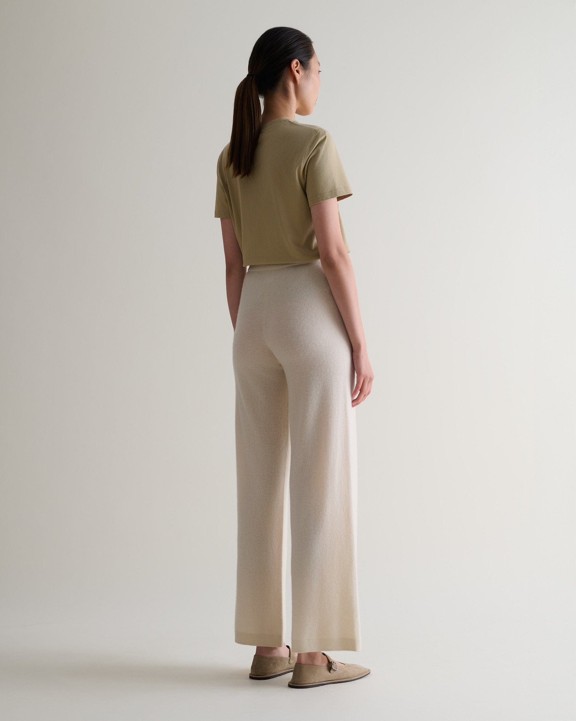Image 3 of 6 showing back of woman wearing Rise & Fall's cream fine cashmere wide-leg trousers with beige t-shirt and shoes.