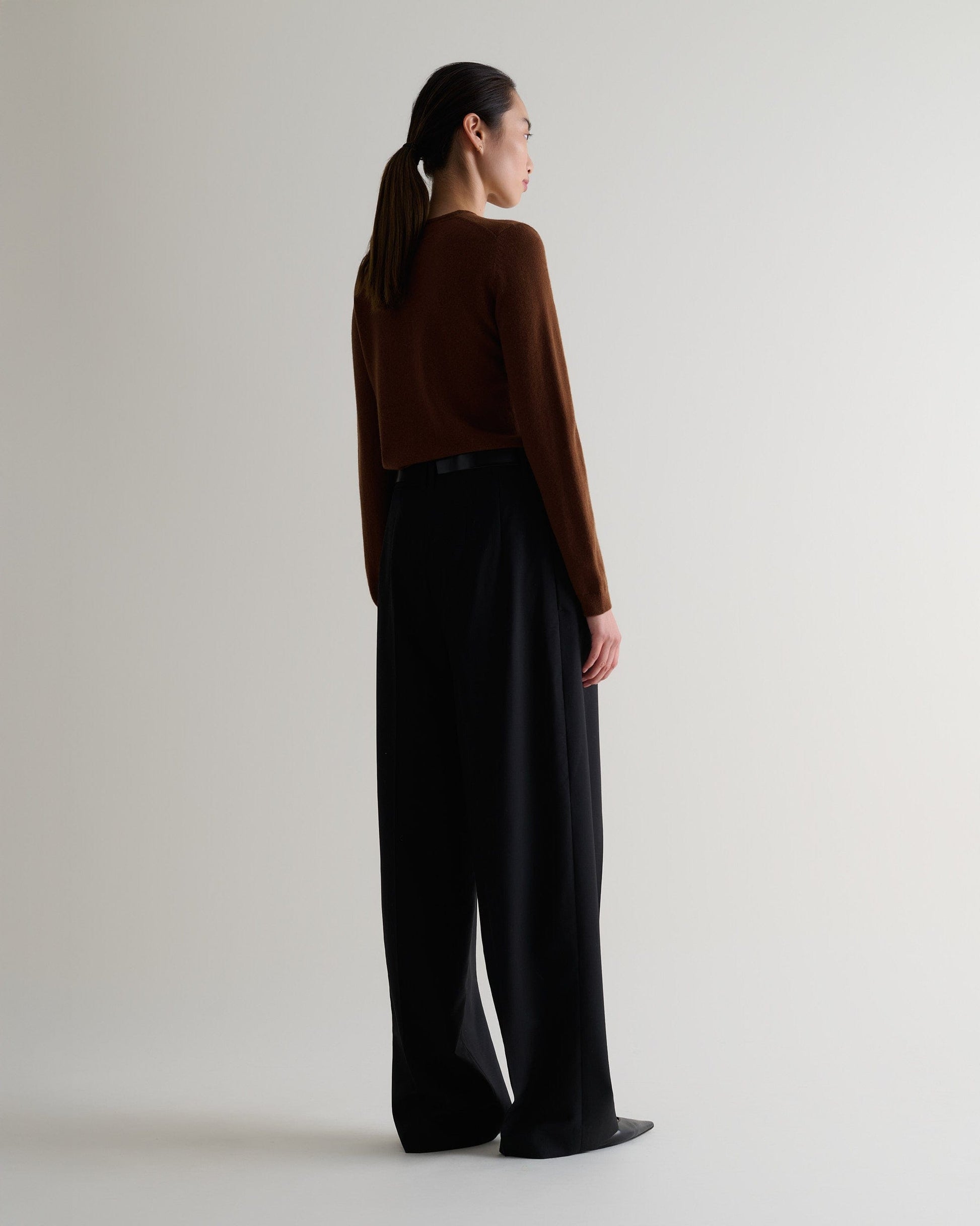 Image 4 of 5 showing back view of a woman wearing Rise & Fall's brown cashmere merino henley jumper with black trousers.