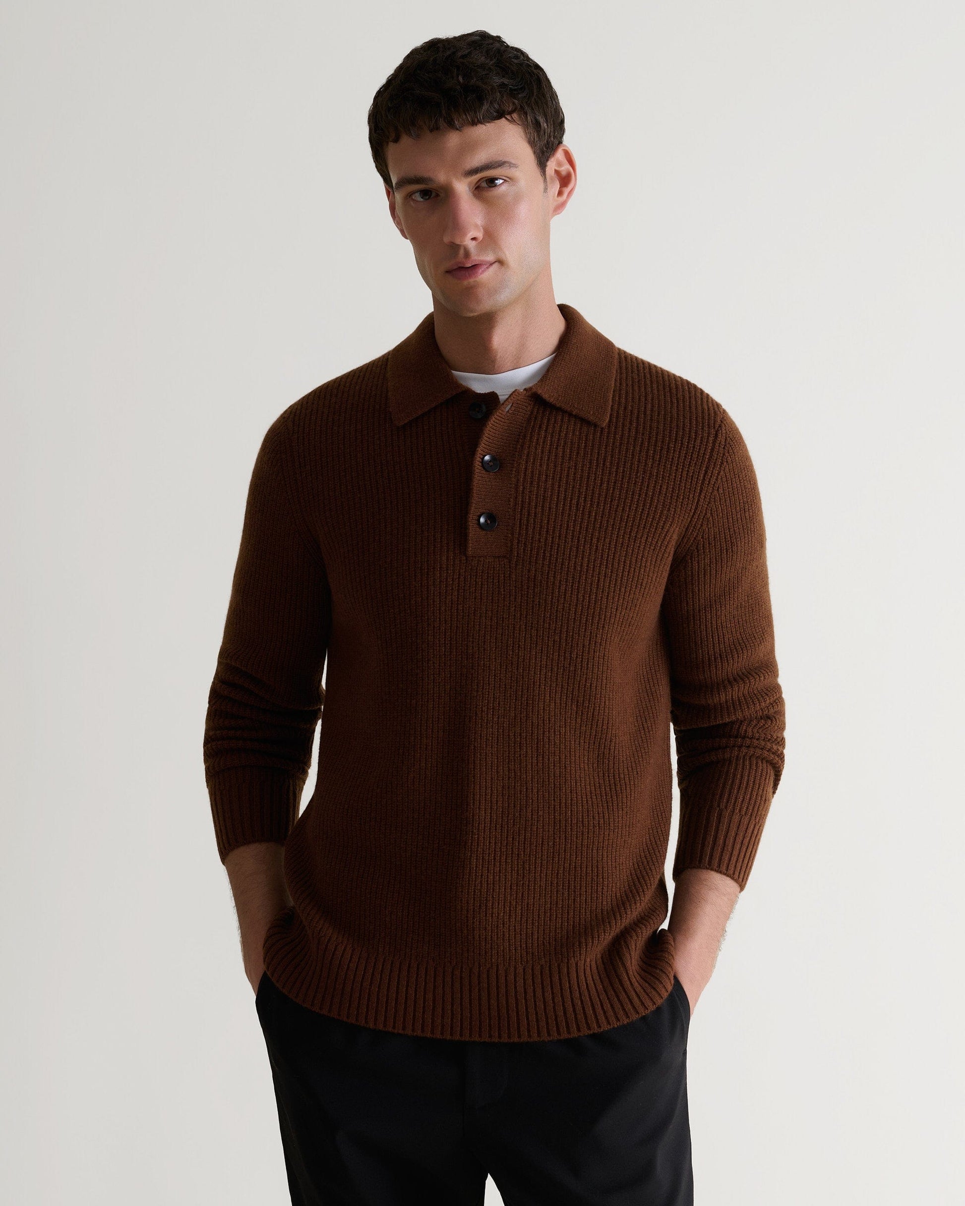 Image 4 of 6 of man wearing Rise & Fall's brown cashmere merino fisherman ribbed buttoned polo with white T-shirt and black trousers.