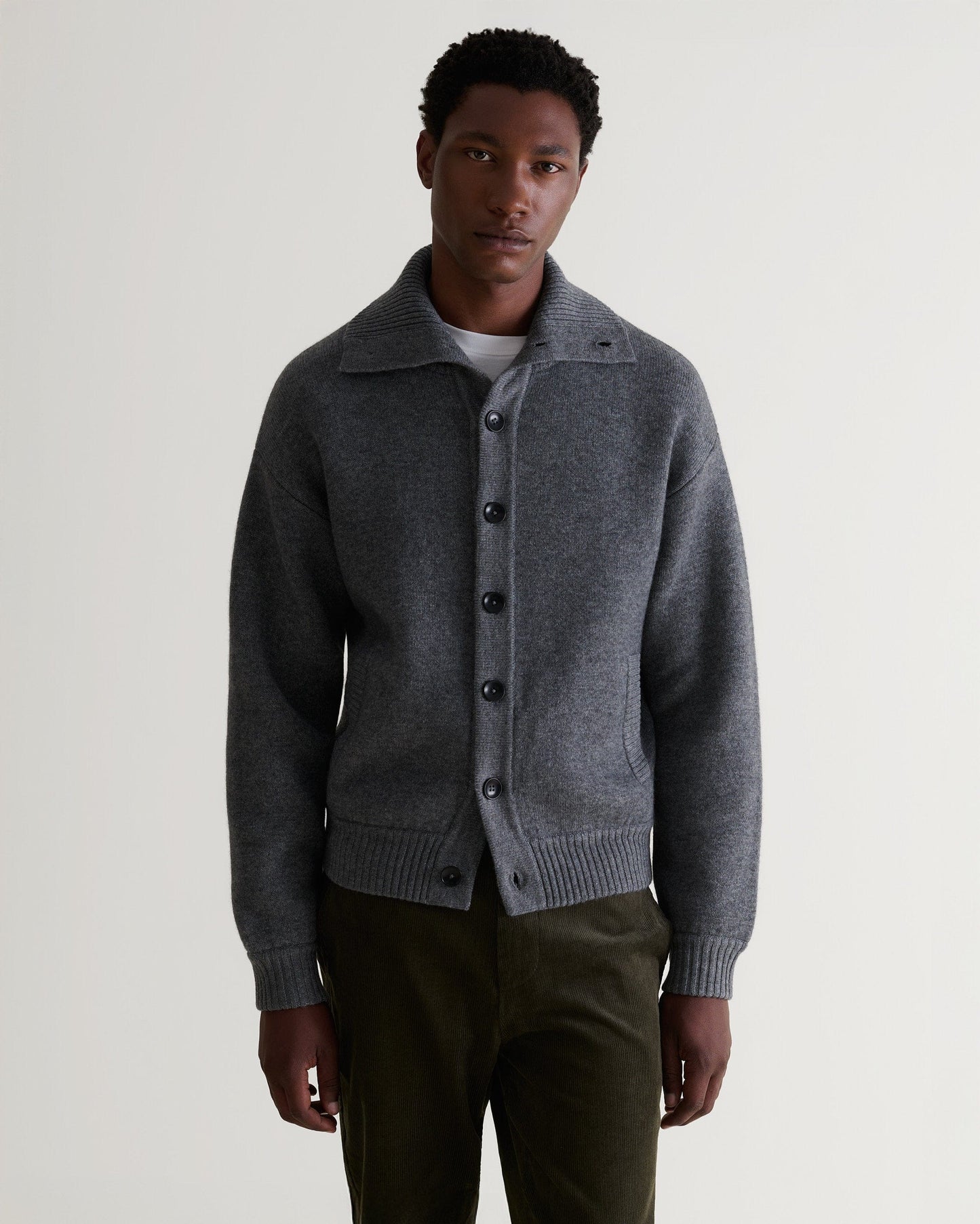 Image 4 of 6 showing man wearing Rise & Fall's dark grey cashmere merino buttoned knitted bomber with white T-shirt and green corduroy trousers.