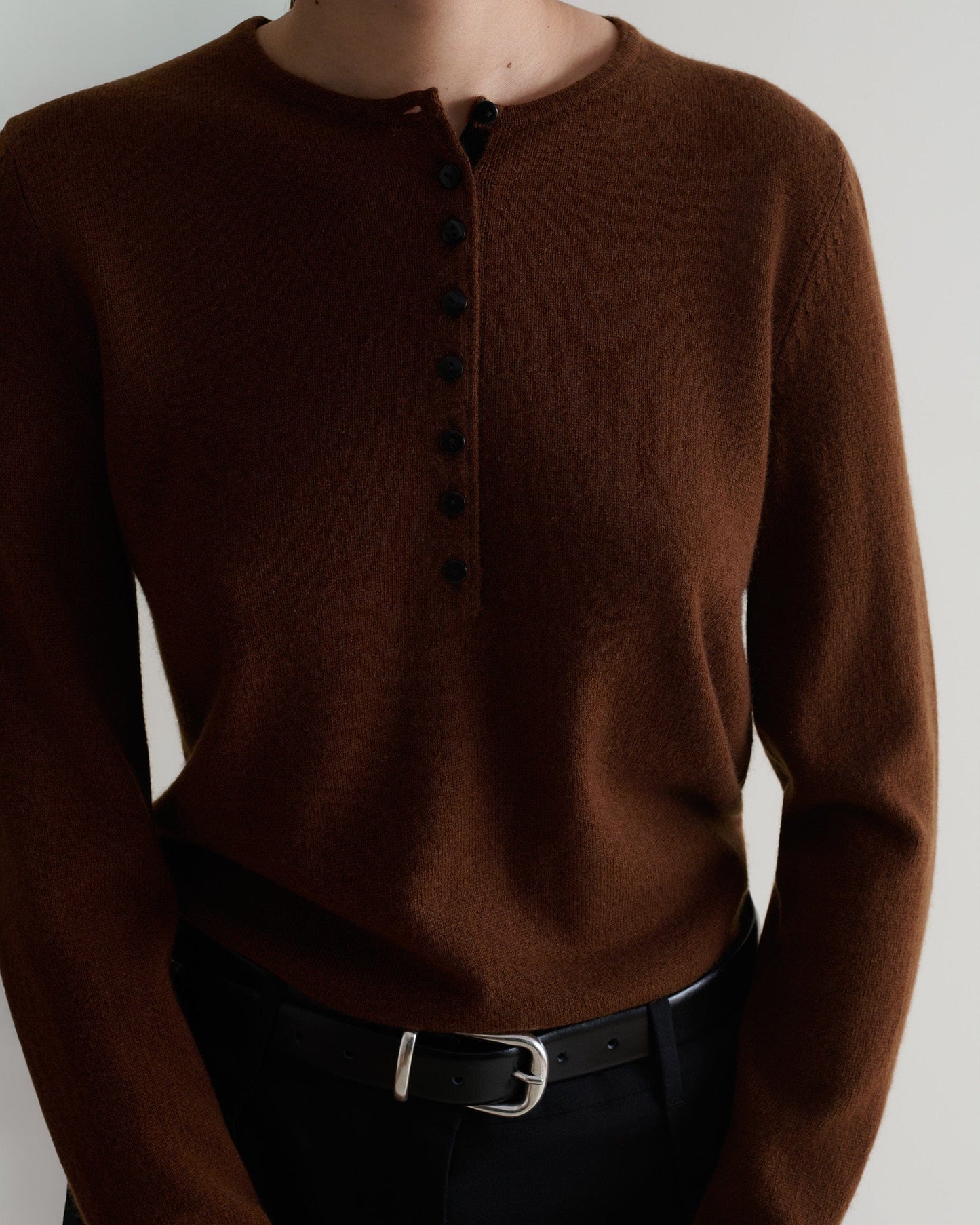 Image 5 of 5 showing a  close up of a woman wearing Rise & Fall's brown cashmere merino henley jumper with black trousers and a black belt. 