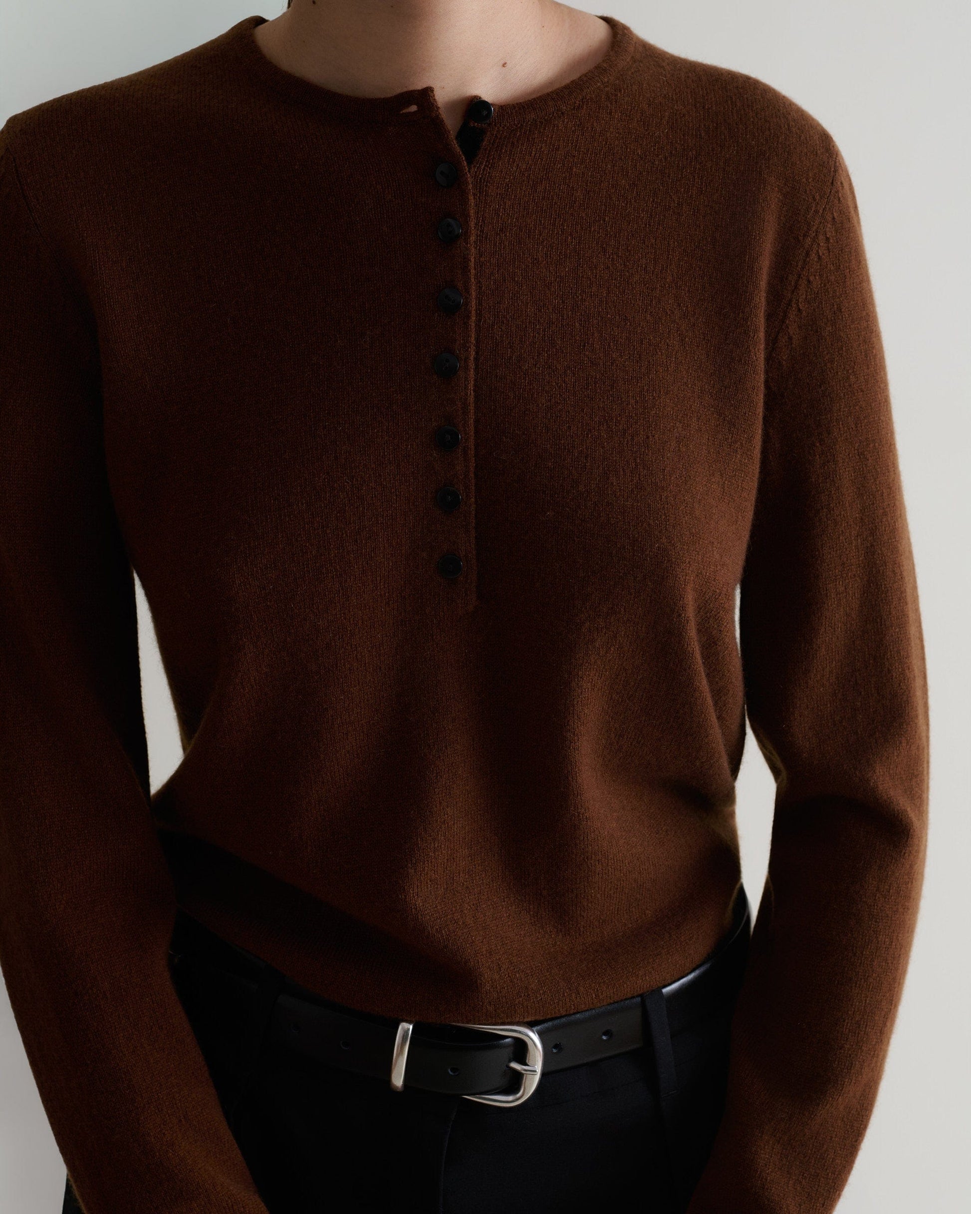 Image 5 of 5 showing a  close up of a woman wearing Rise & Fall's brown cashmere merino henley jumper with black trousers and a black belt. 