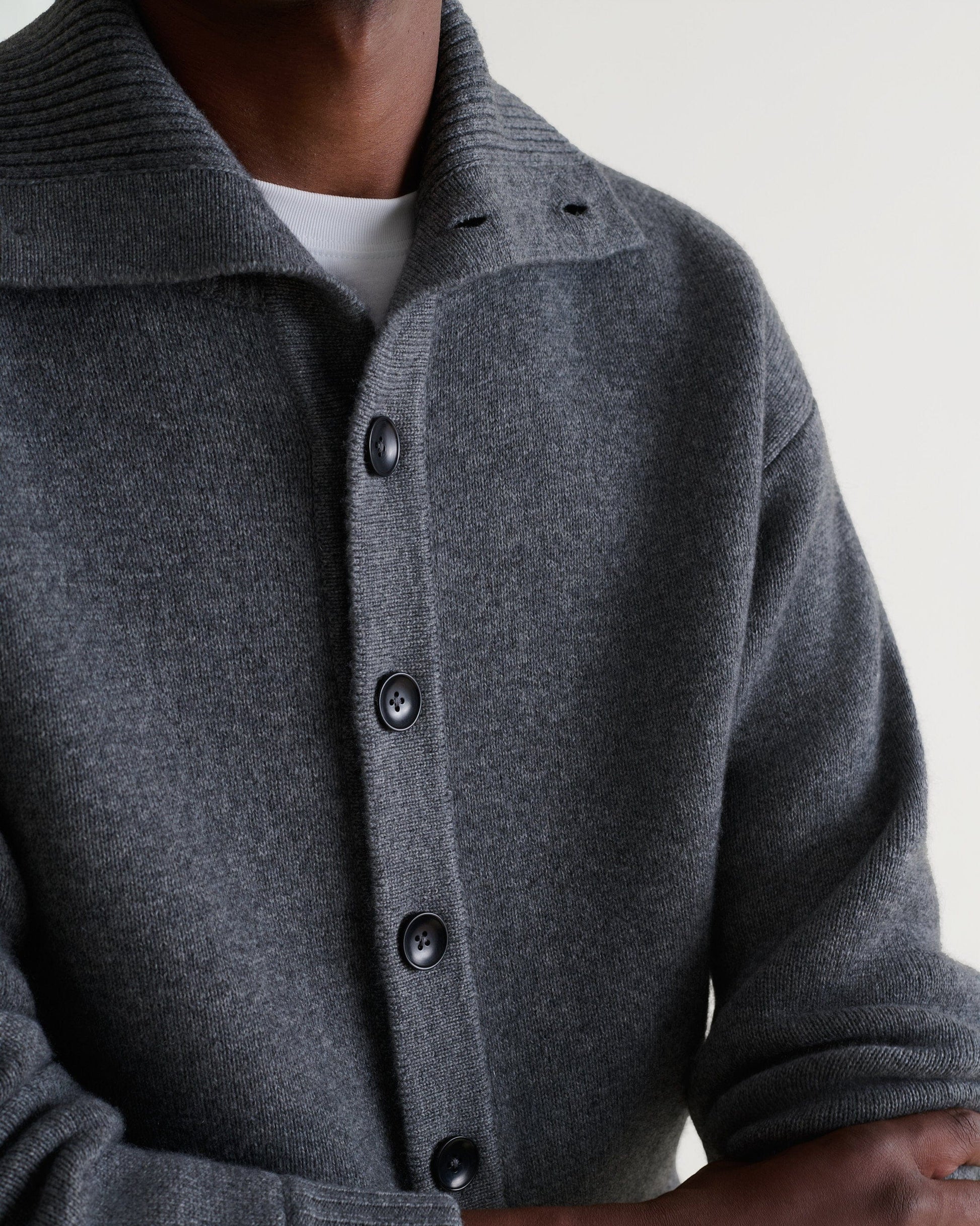 Image 5 of 6 showing close up of man wearing Rise & Fall's dark grey cashmere merino buttoned knitted bomber. Worn with white T-shirt.