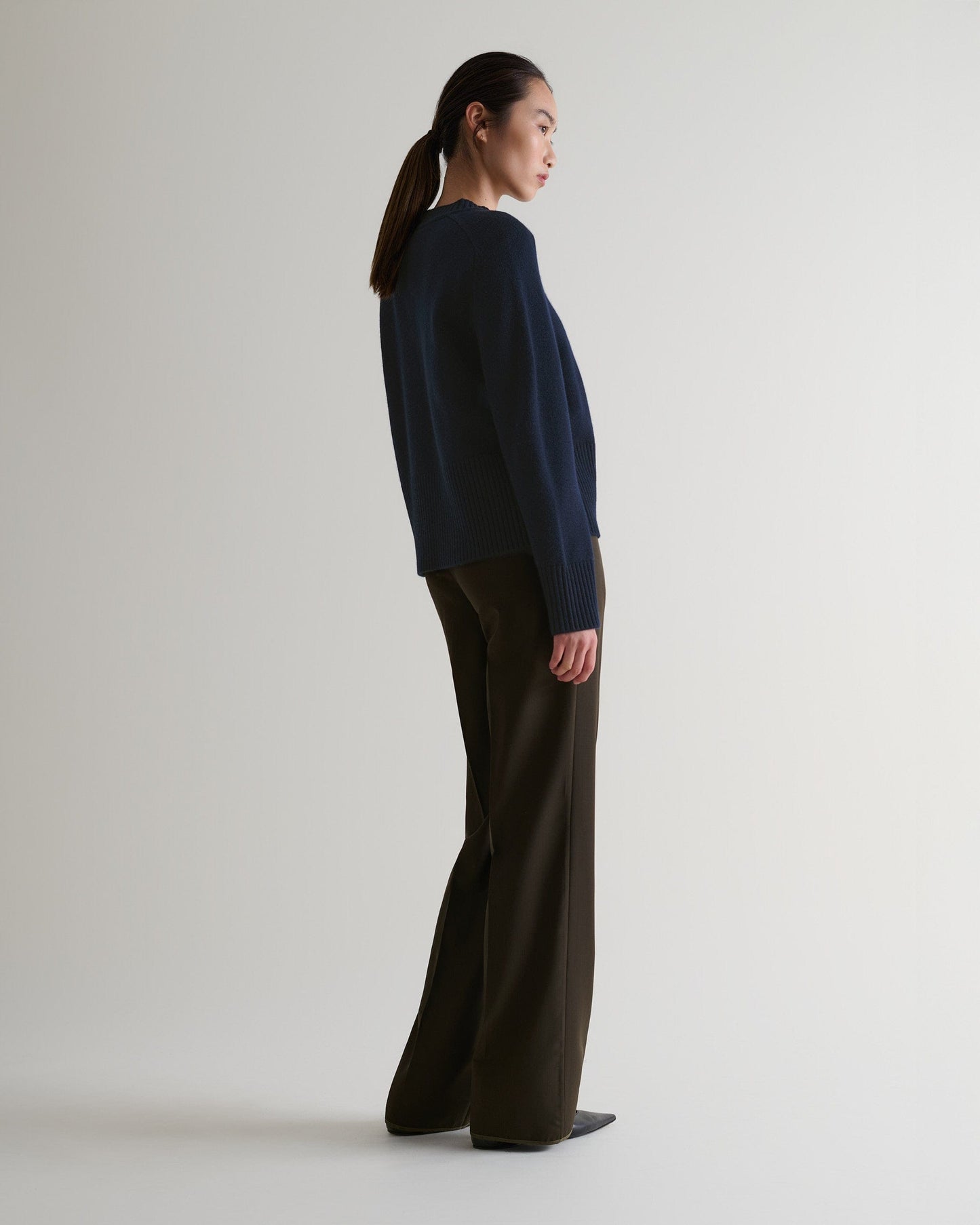 Image 5 of 6 showing side of woman wearing Rise & Fall indigo cashmere merino saddle sleeve crew neck jumper with brown trousers.