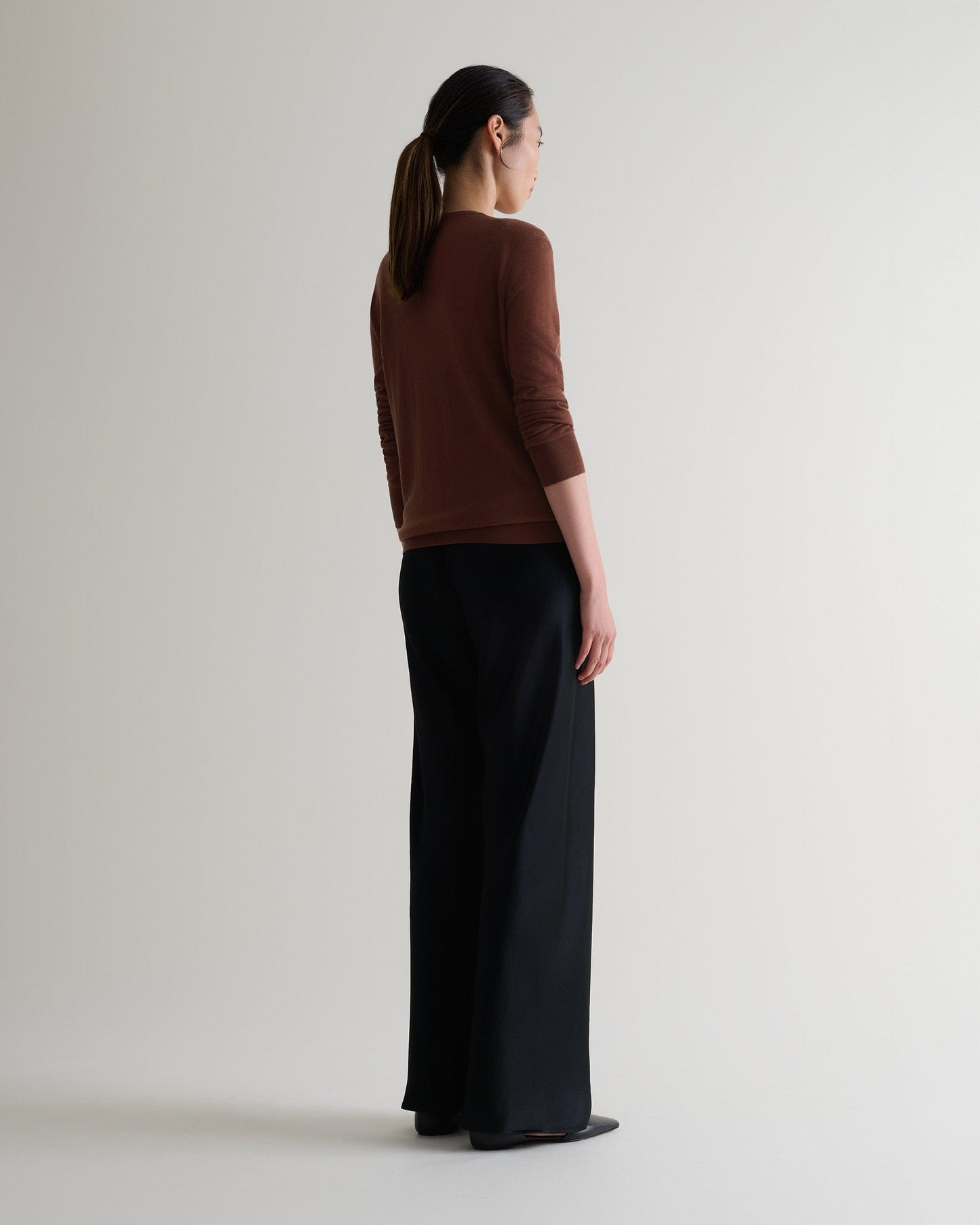 Image 5 of 6 showing full length side view of woman wearing Rise & Fall's brown tencel cashmere double layered crew neck jumper with black trousers. 