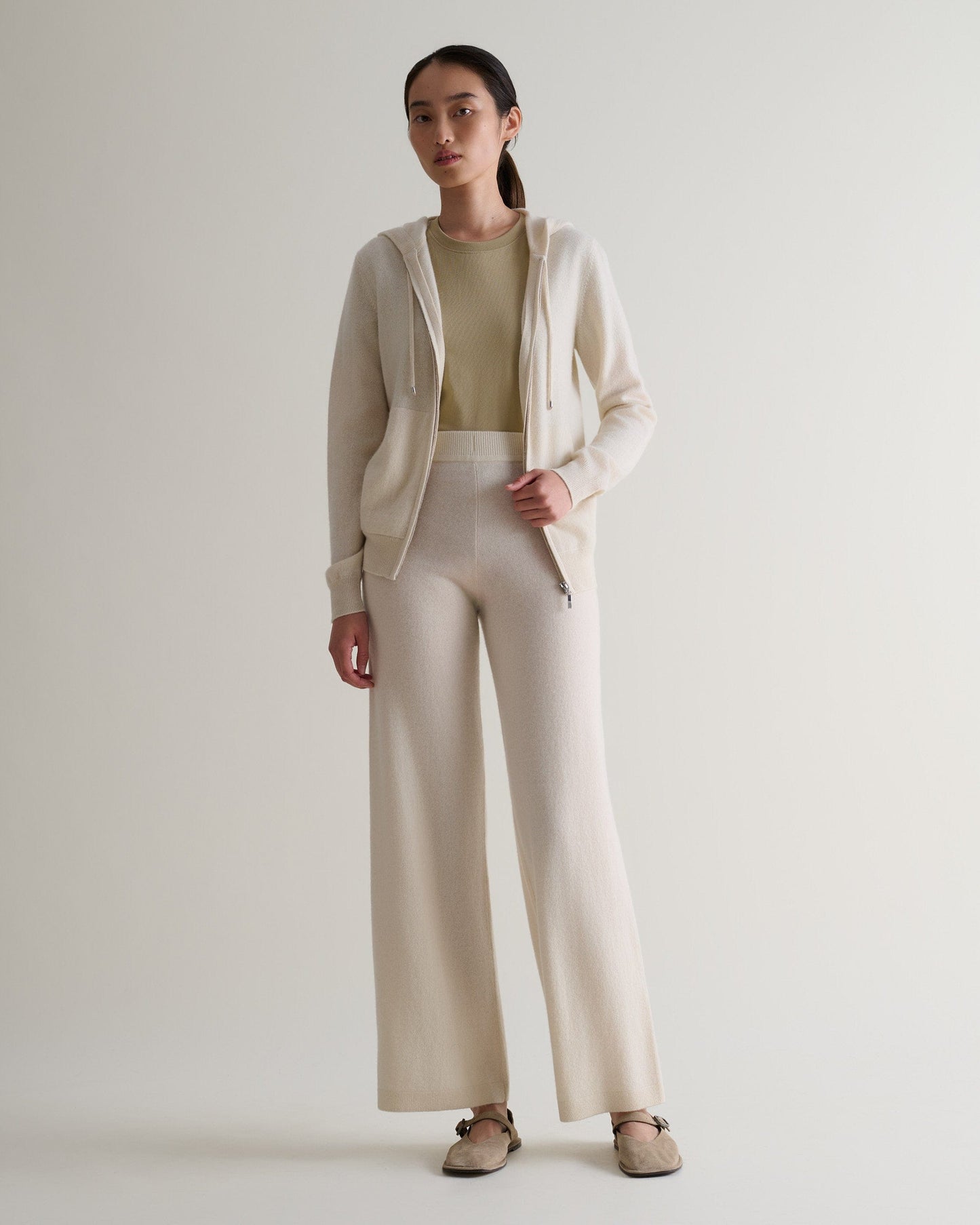 Image 6 of 6 showing woman wearing Rise & Fall's cream fine cashmere wide-leg trousers with matching cashmere hoodie and a beige t-shirt.