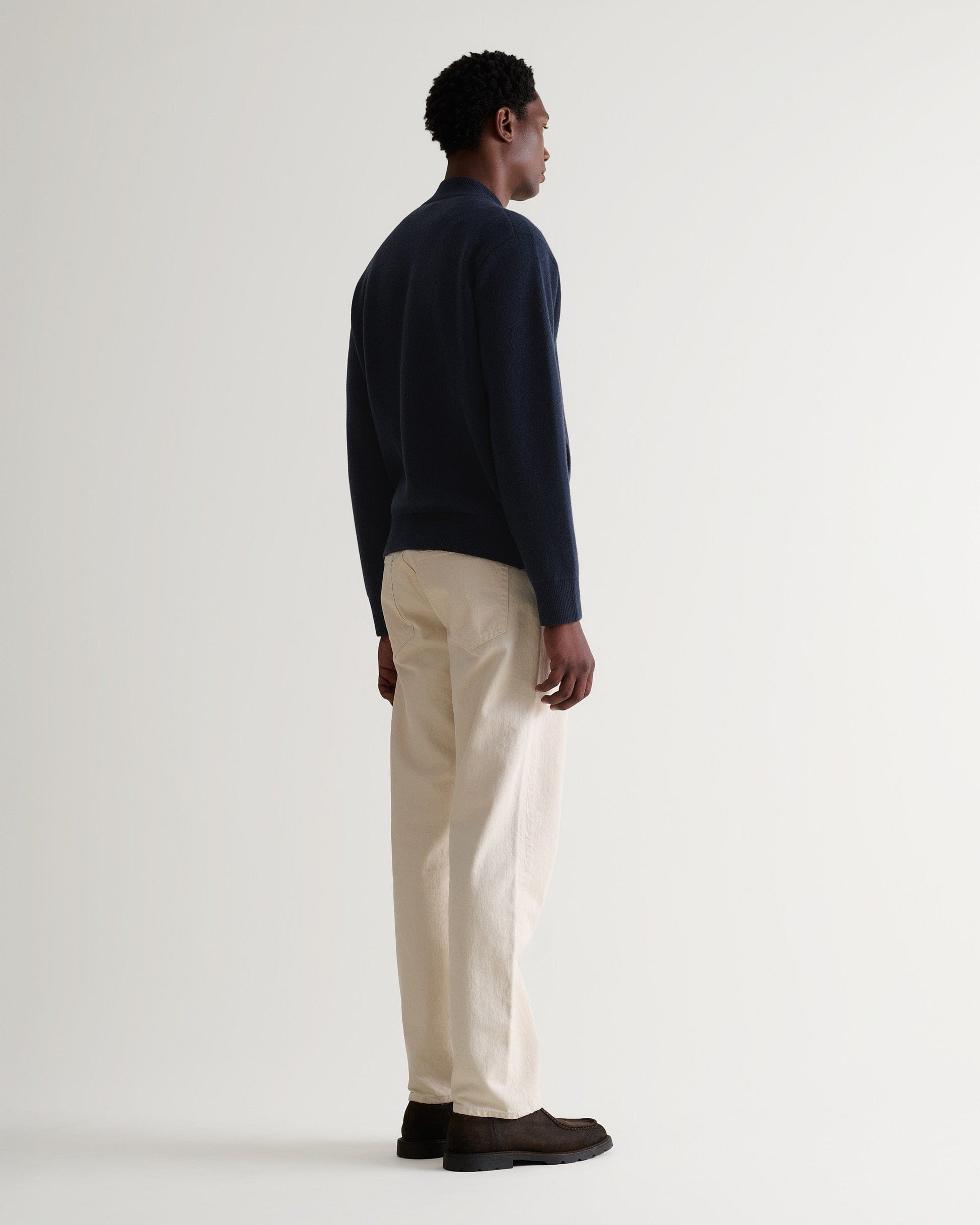 Back view of man wearing navy cashmere merino double-faced knitted bomber with cream trousers and brown shoes.