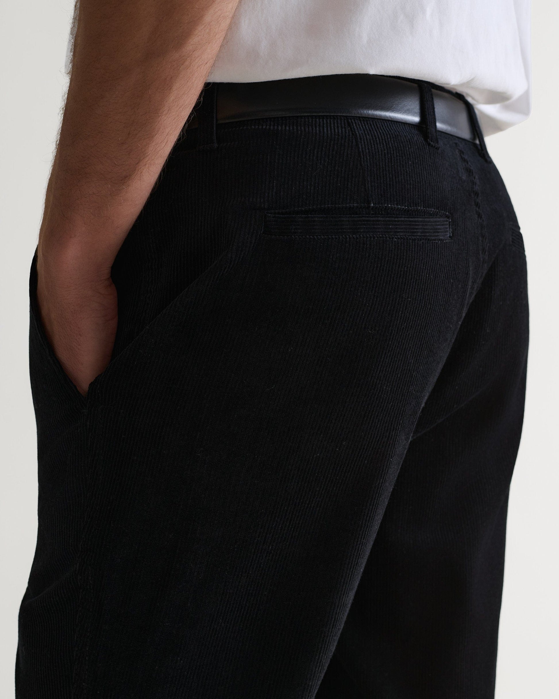 Close up back view of man wearing Rise & Fall's black corduroy trousers with black leather belt and white T-shirt.