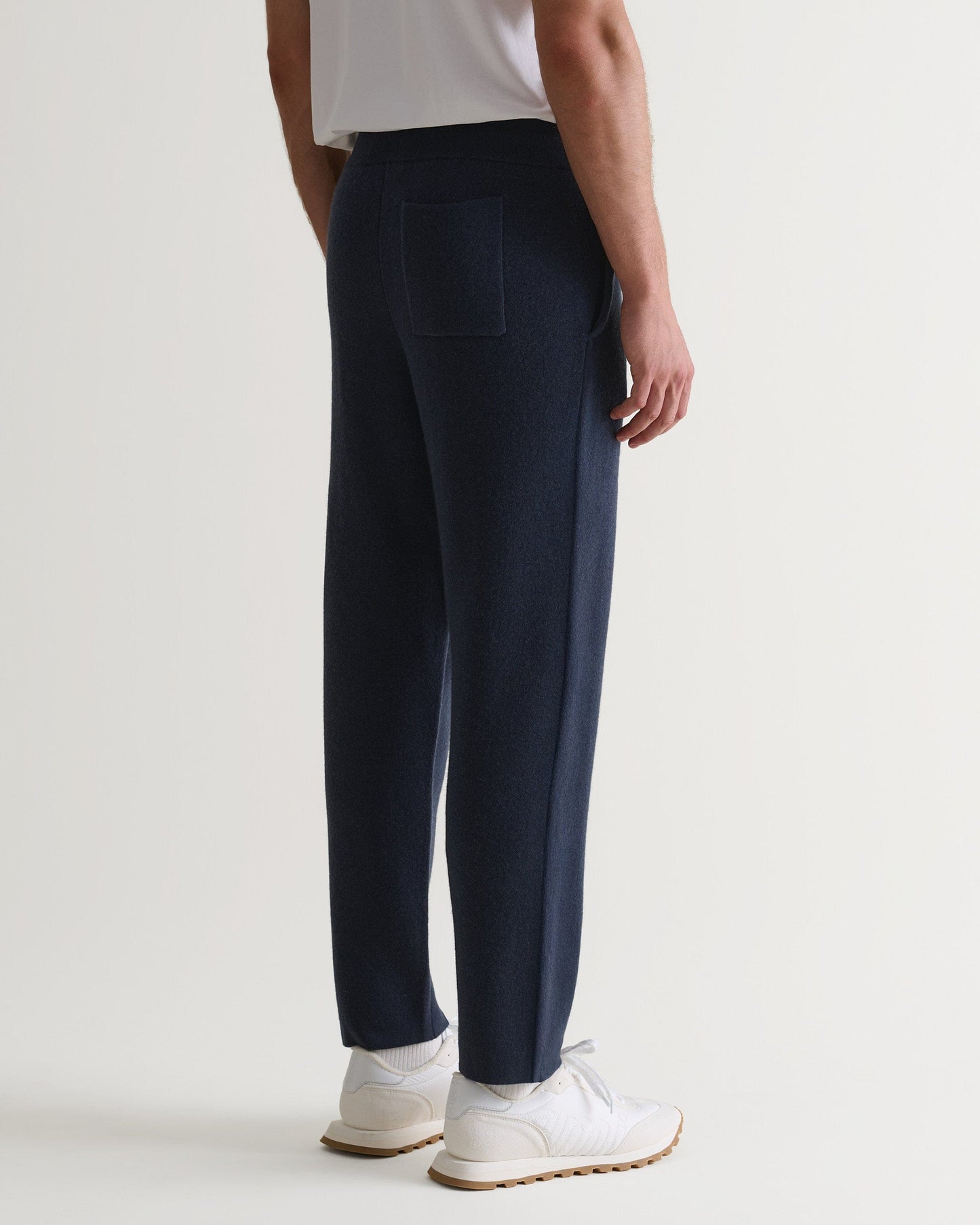 bottom half side view of man wearing Rise & Fall's navy cashmere merino double-faced joggers with white T-shirt and trainers.