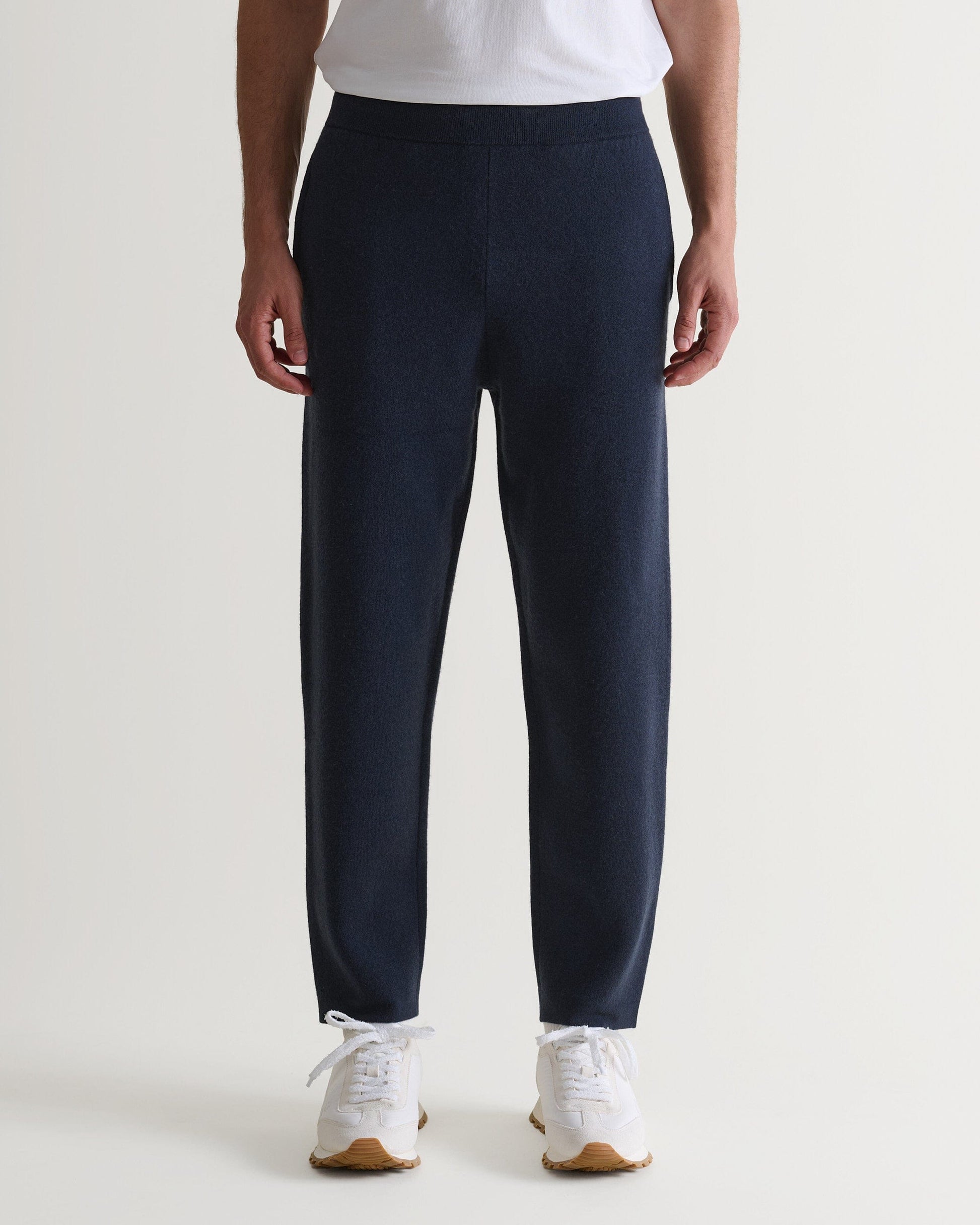Bottom half of man wearing Rise & Fall's navy cashmere merino double-faced joggers with white T-shirt and trainers. 