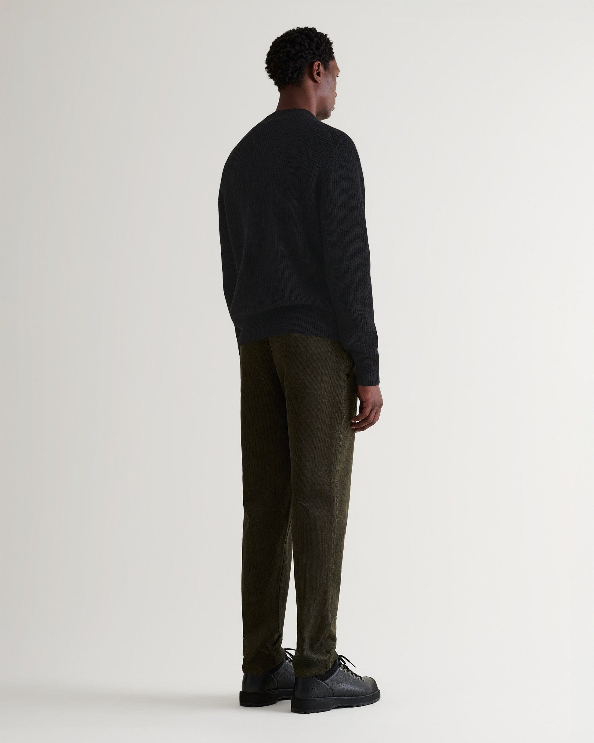 Man wears black cashmere merino stand neck ribbed jumper with green corduroys and black shoes. Full length back view.