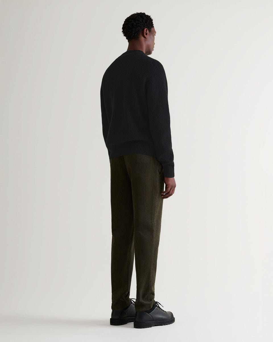 Man wears black cashmere merino stand neck ribbed jumper with green corduroys and black shoes. Full length back view.