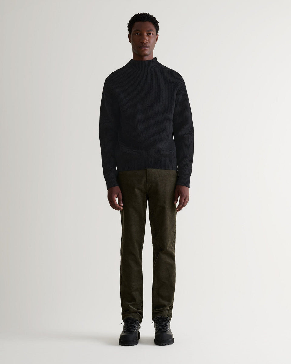 Full length front view of man wearing black cashmere merino stand neck ribbed jumper with green corduroys and black shoes.
