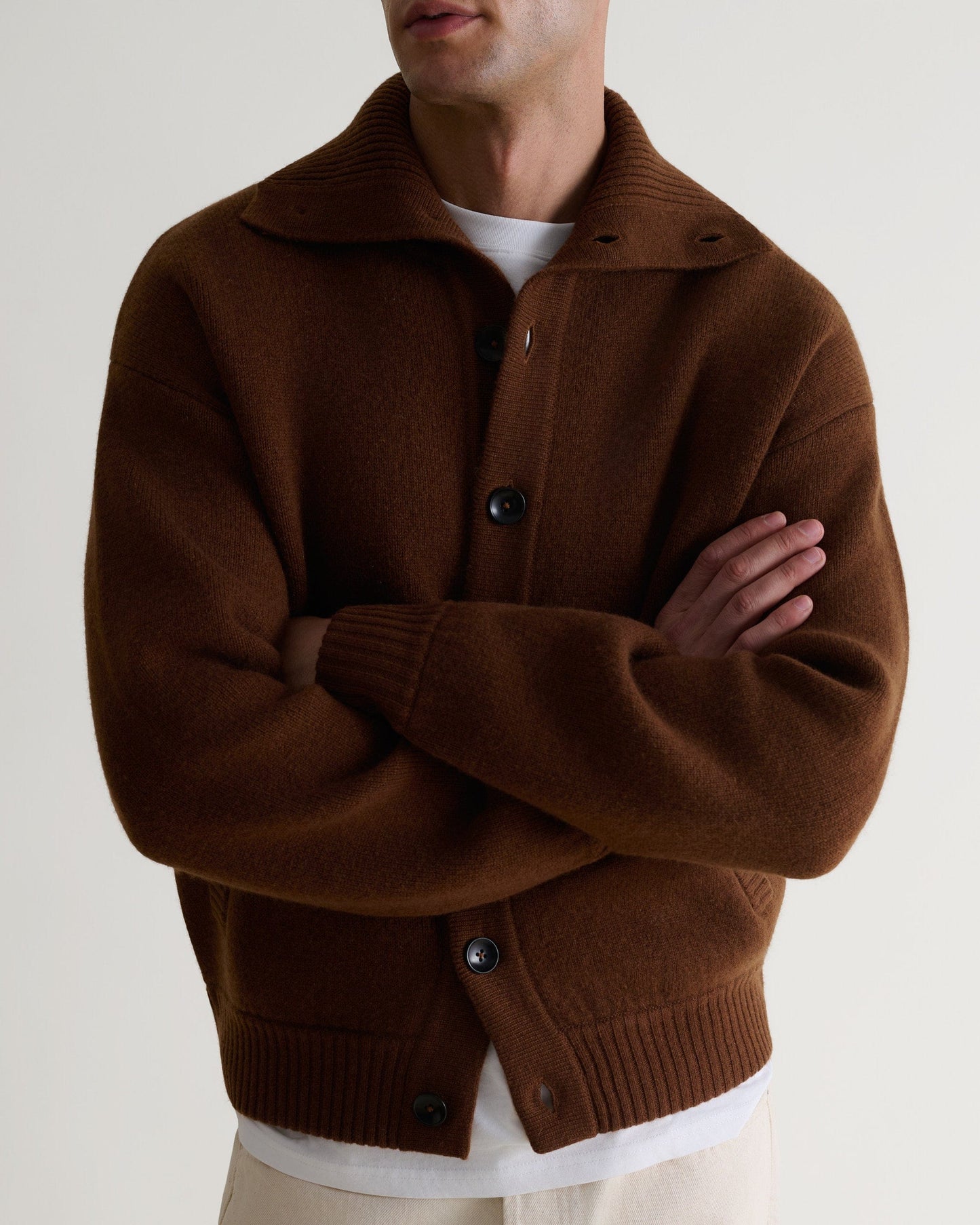 Image 5 of 6 showing close up of man wearing Rise & Fall's burl brown cashmere merino buttoned knitted bomber. Worn with white T-shirt and cream trousers.