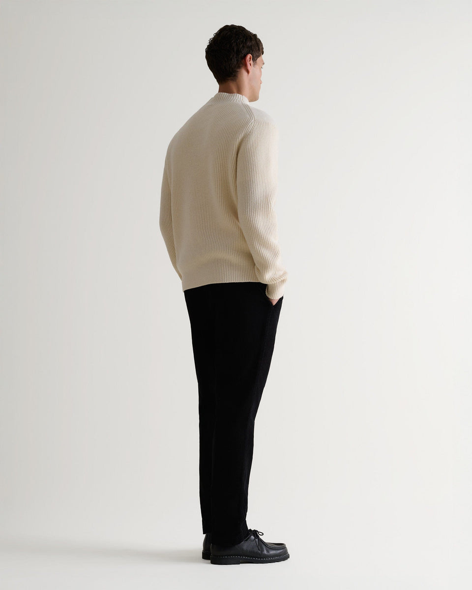 Full length side view of man wearing cream cashmere merino stand neck ribbed jumper with black trousers and shoes.