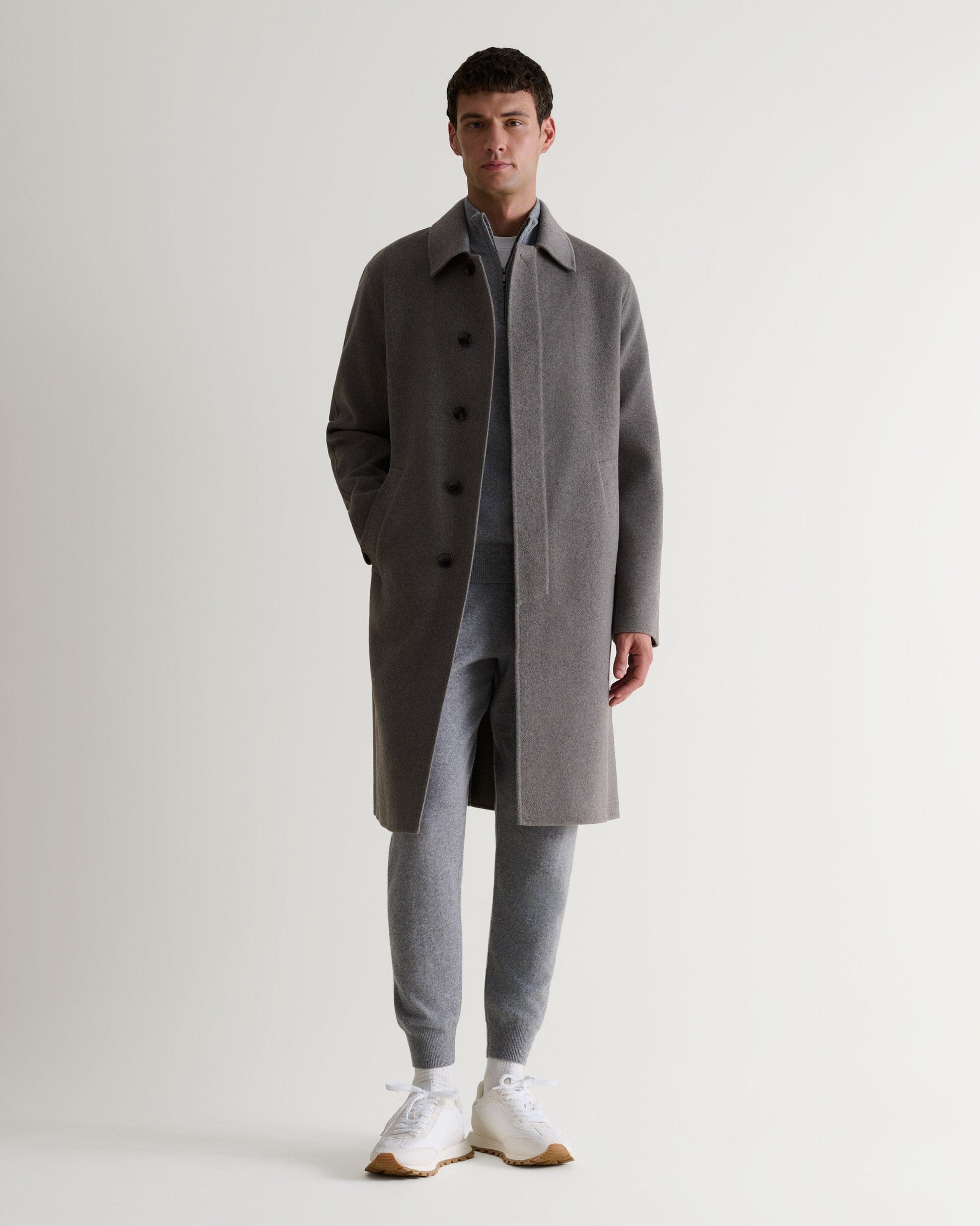 Man wears fawn cashmere merino double-faced wool coat. Matched with grey cashmere jumper and joggers. Full length view.